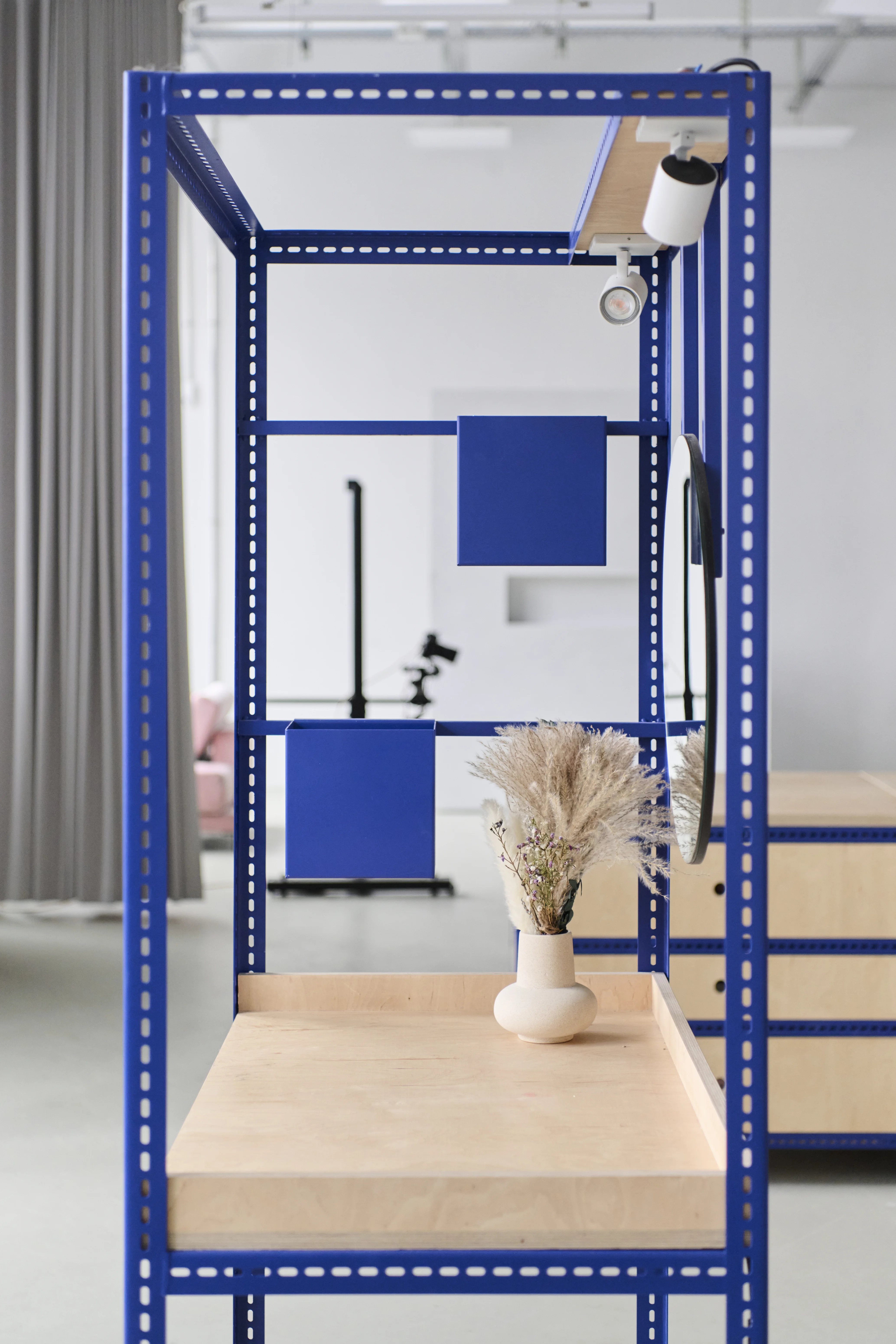Blue steel-frame studio module at the FLACONI CONTENT STUDIO with a birch worktop, dried flowers in a white vase, mounted spotlight and blue metal panels.