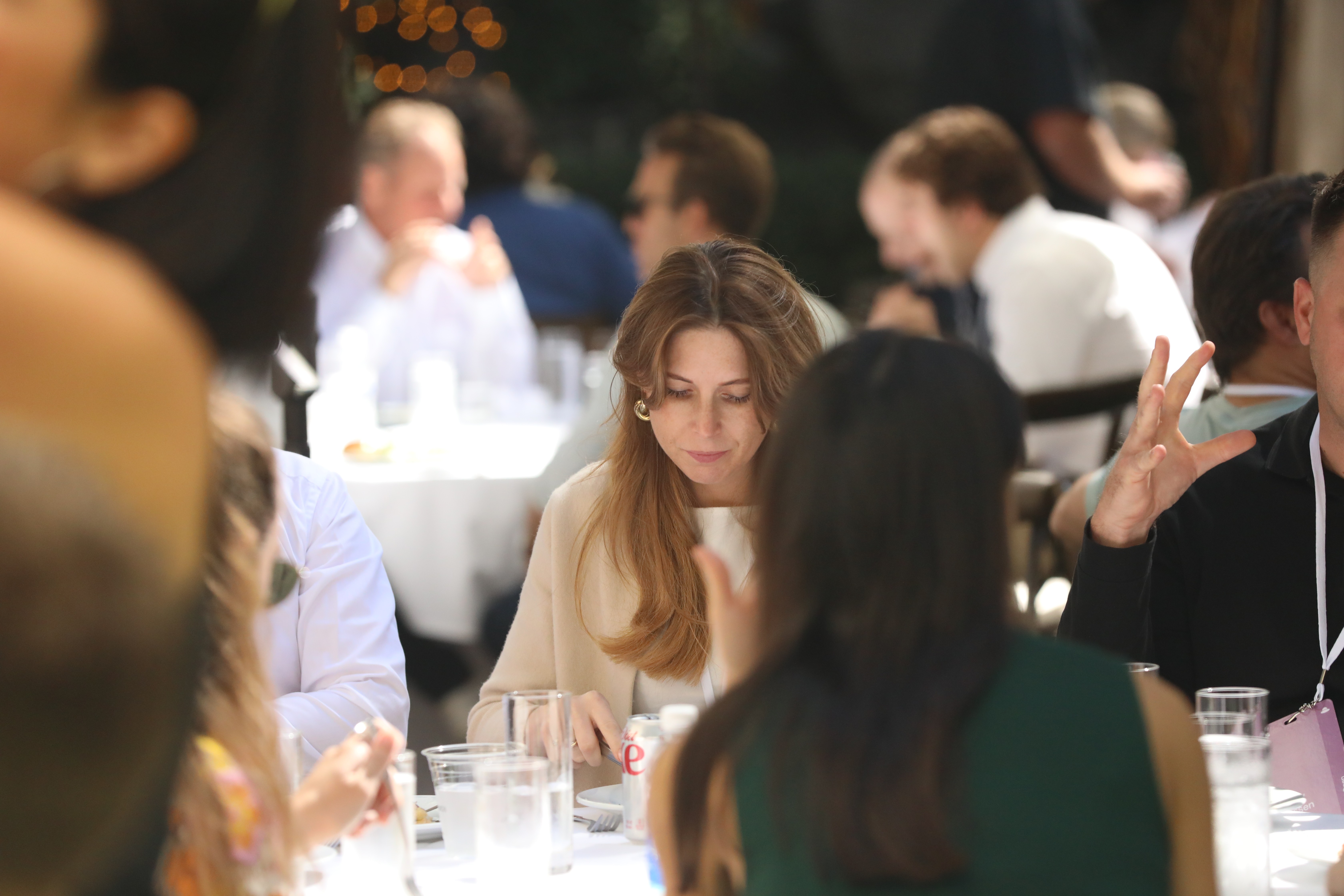 Professionals gathered at a networking lunch, with a woman in a beige top in focus at center while colleagues converse at surrounding tables.