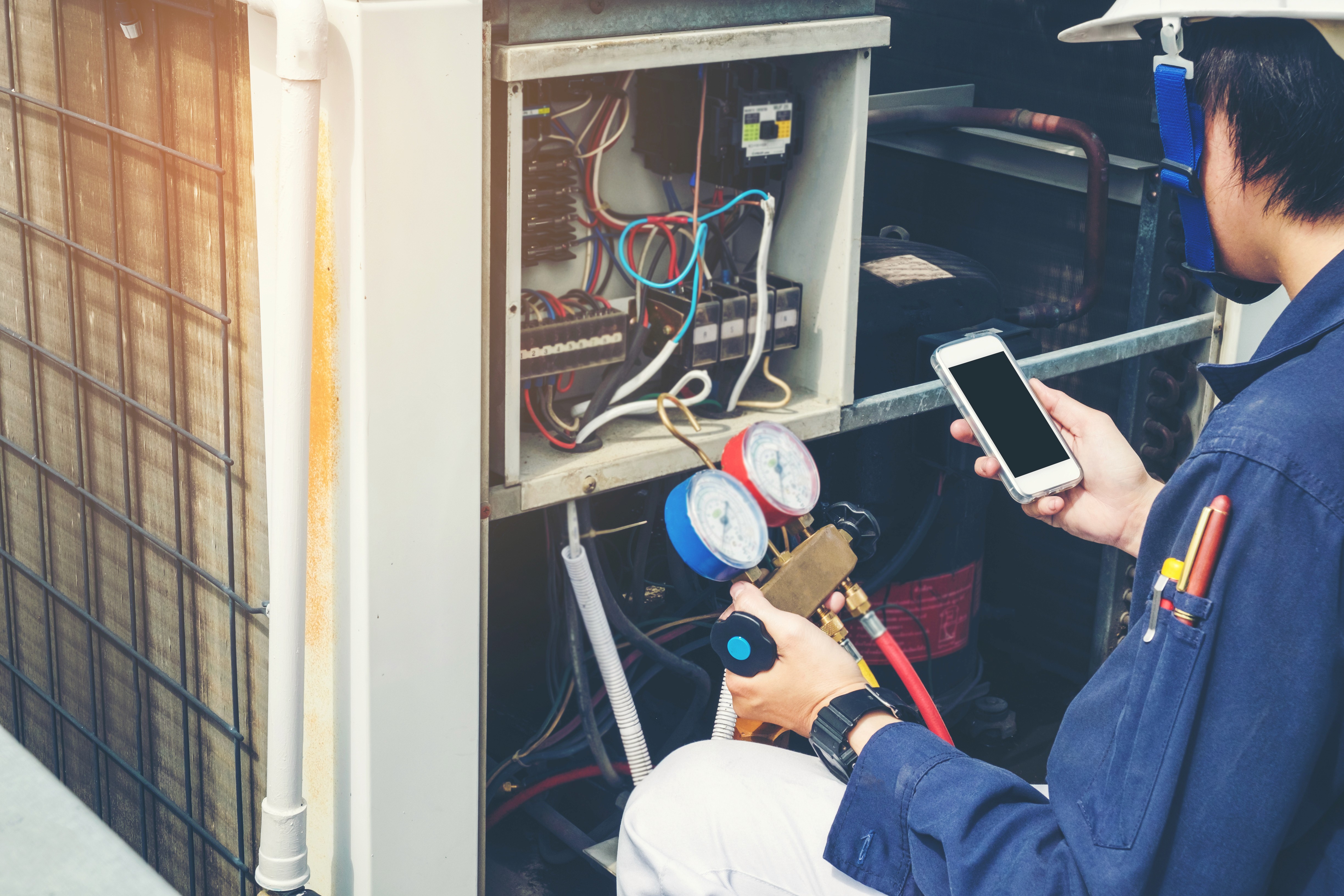 A technician in a blue uniform, holding a smartphone and pressure gauges, is performing maintenance on an open air conditioning unit, with visible wires and components, highlighting HVAC repair and diagnostics.