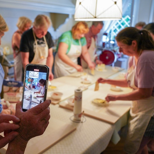 People in aprons preparing food around a table in a kitchen while someone takes a photo with a smartphone.