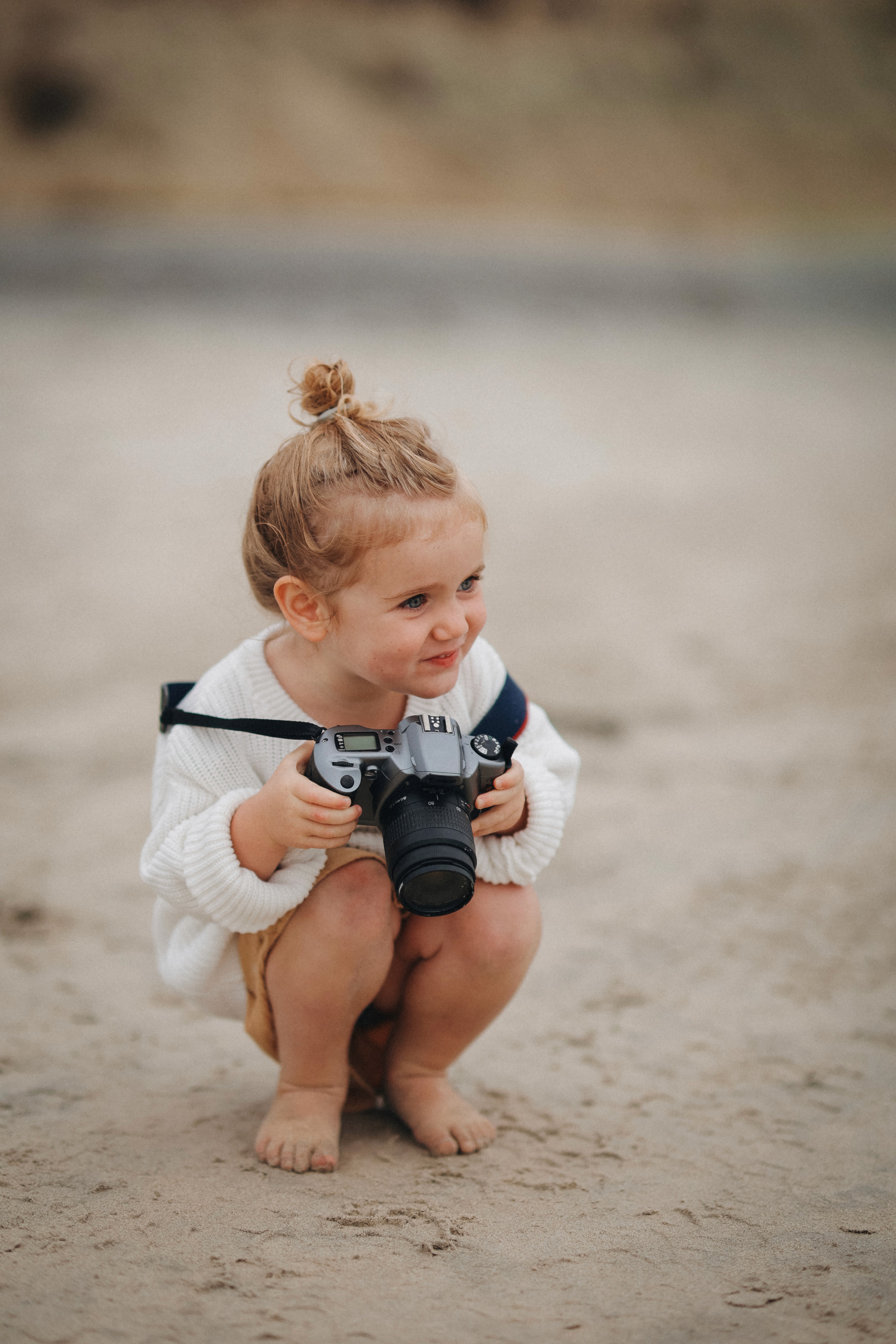 Little girl crouching on the sand holding a camera during a family photo session