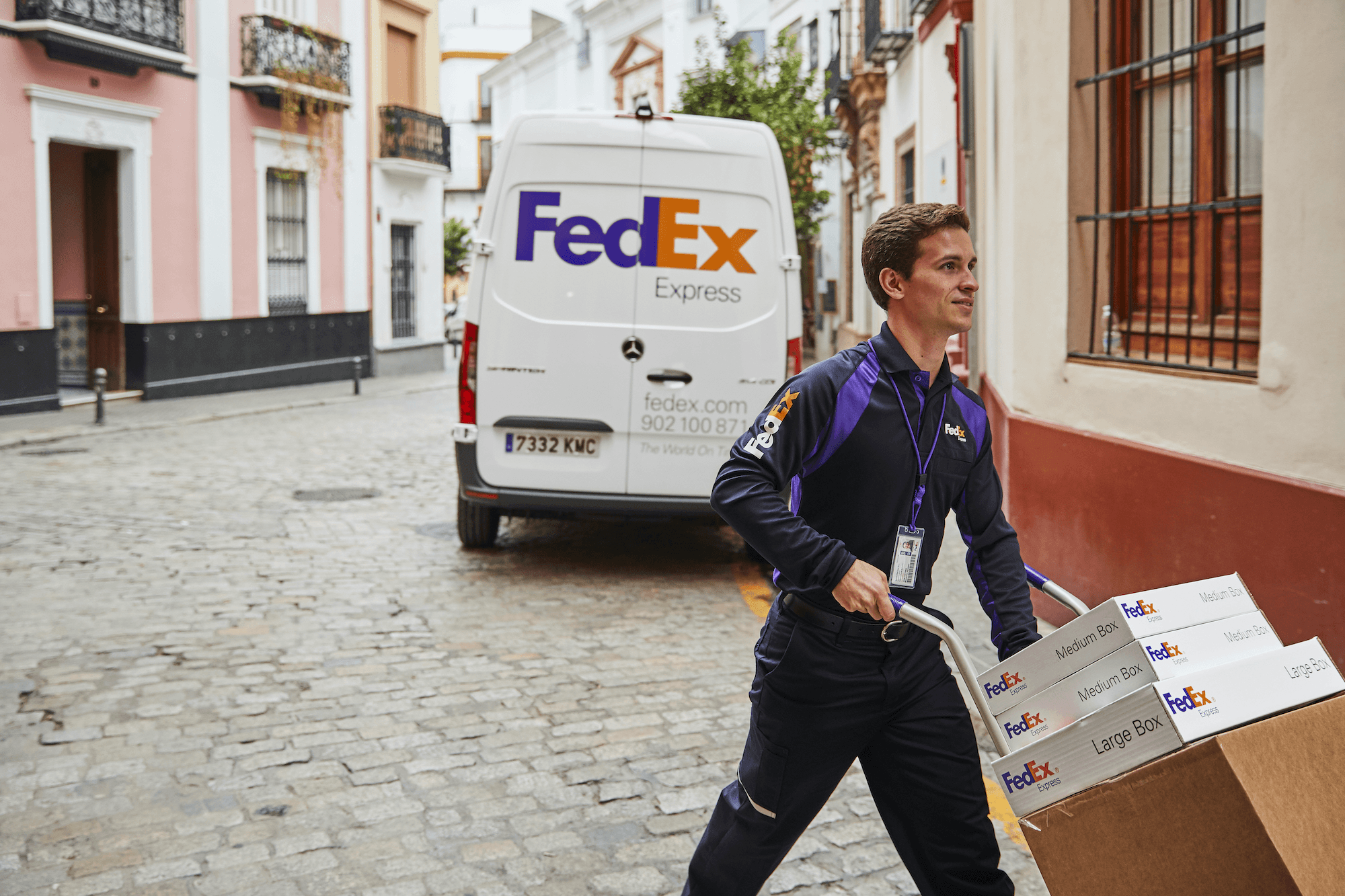 Fedex Postbote, der Sackkarre mit Paketen durch Straße schiebt mit Fedex Transportvan im Hintergrund