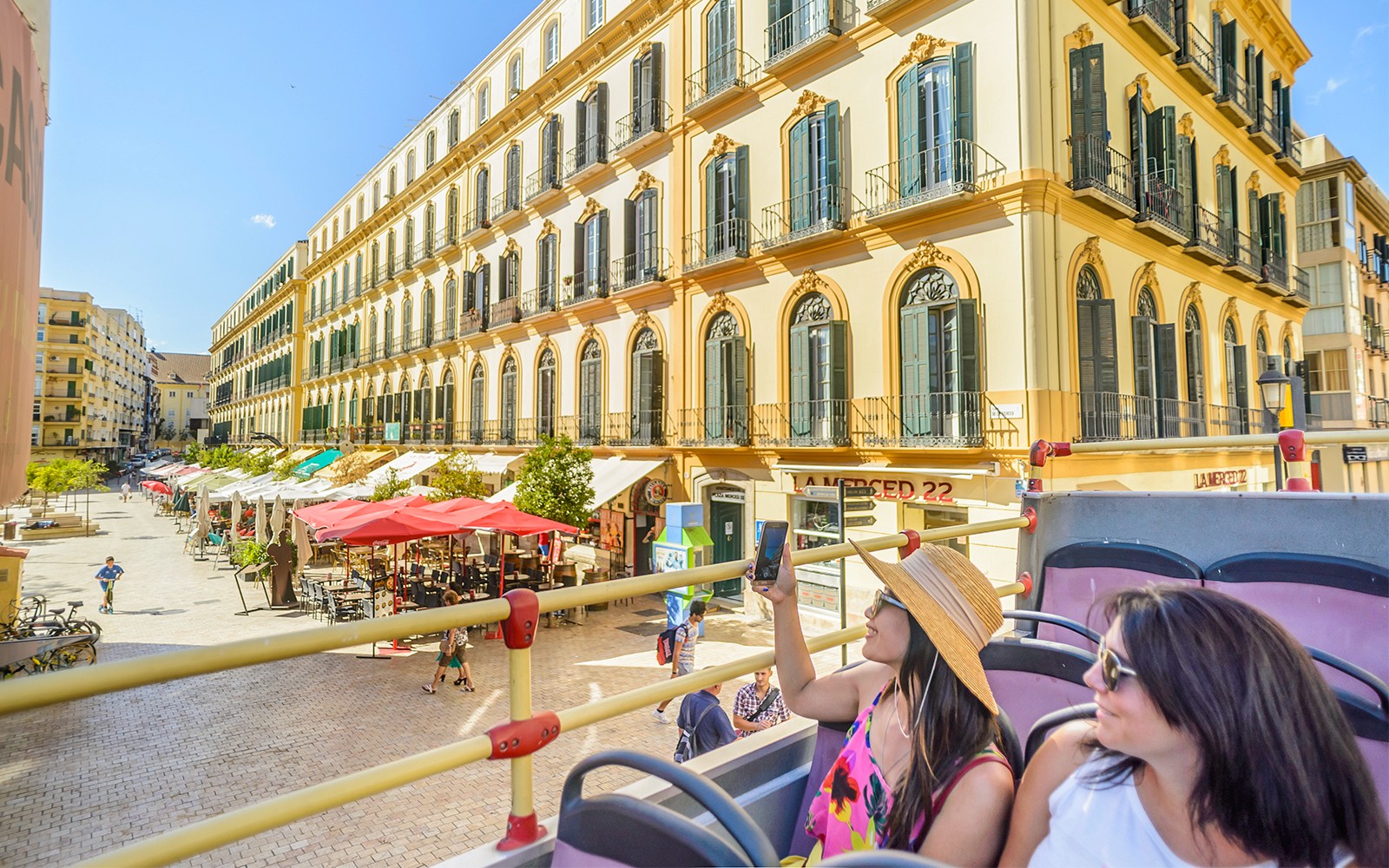 Passengers on Malaga hop-on hop-off tour bus passing historic buildings.