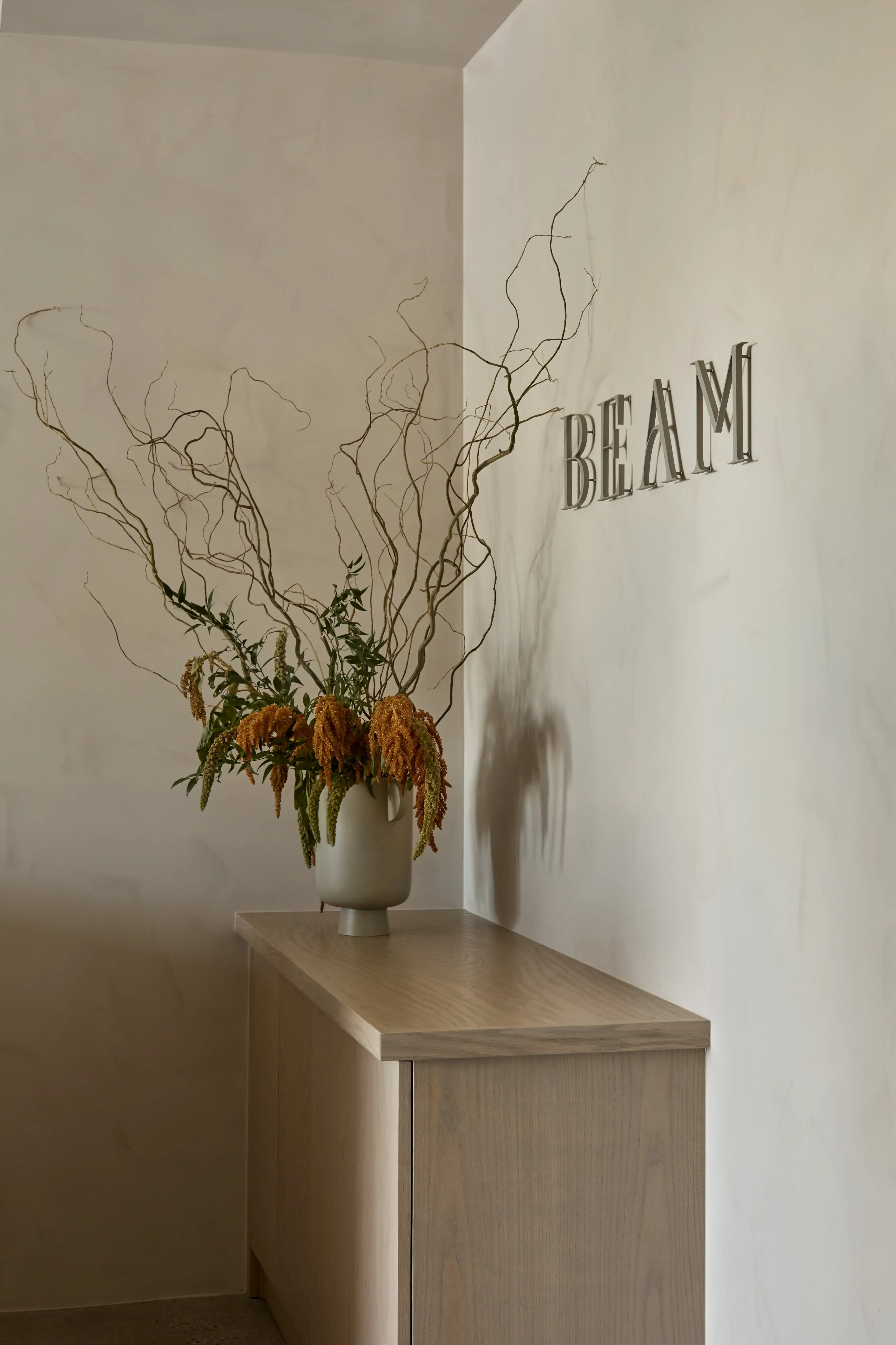 Close-up of BEAM metal wall signage mounted above a custom light wood reception desk in a modern wellness studio.