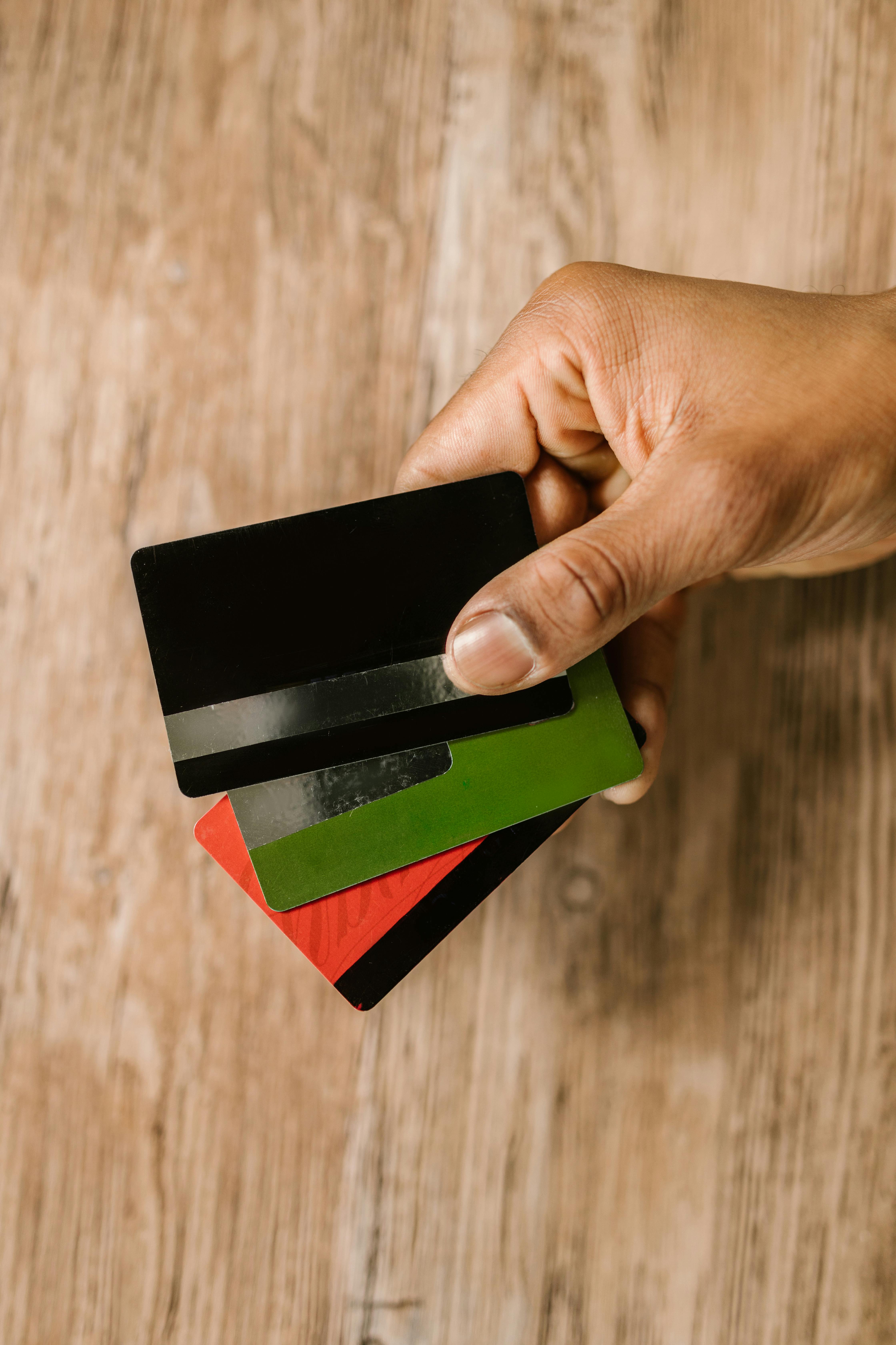 A hand holds three credit cards, one black, one green, and one red, over a wooden surface, showcasing a variety of card options for financial transactions.