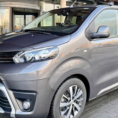 Minivan gris estacionada, mostrando parte frontal y lateral, con luces diurnas encendidas. La entrada de un edificio es visible al fondo.