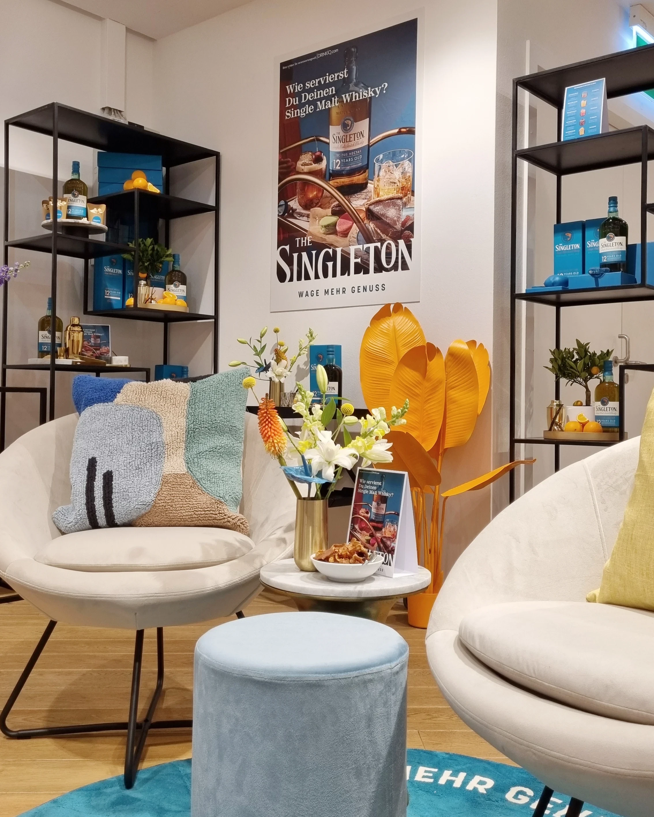 Lounge with cream armchairs, velvet pouf, flowers and Singleton poster between display shelves.
