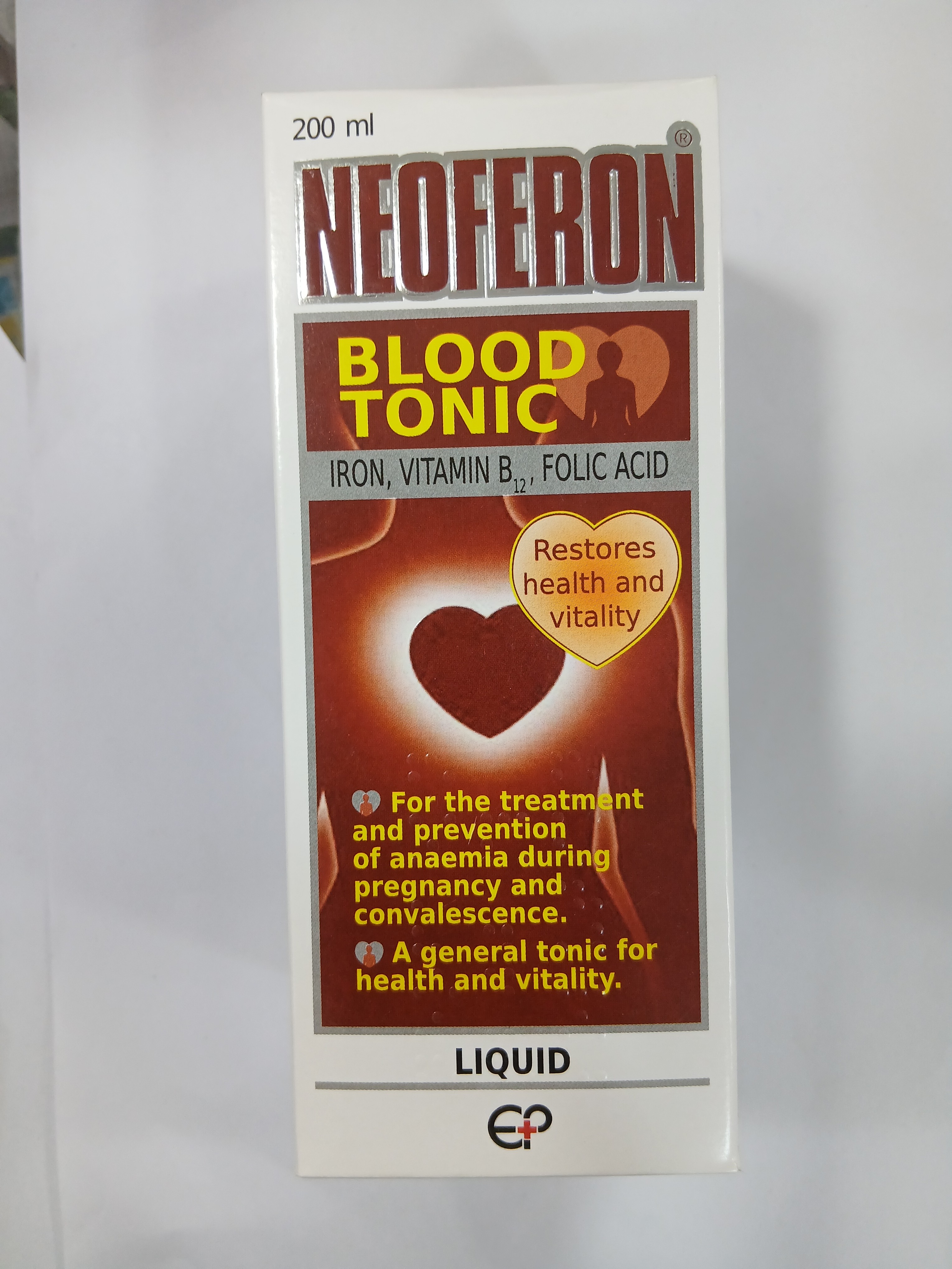 White cardboard packaging for a liquid supplement displays a red and orange label with text indicating it contains iron, vitamin B, and folic acid, and features a heart graphic alongside claims of restoring health and vitality.
