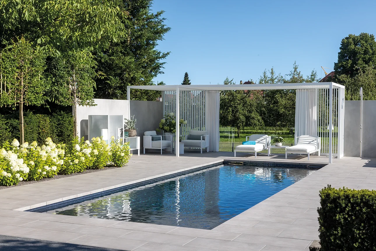 MLine Rechteckpool mit weißer Pergola, weißen Loungemöbeln und blühenden Gartensträuchern