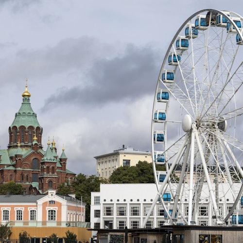 A Ferris wheel with blue gondolas stands in the foreground, while a red-brick church with green domes is visible in the background.