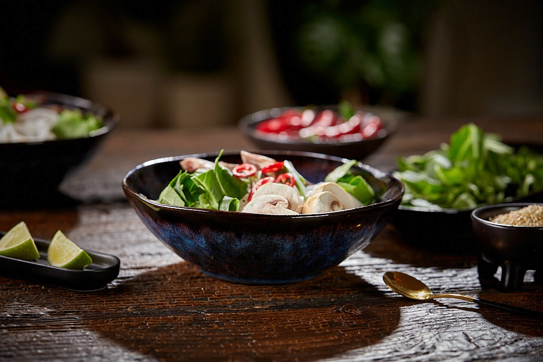 A rustic wooden table features a dark ceramic bowl filled with a fresh, colorful salad, including sliced mushrooms, chili, and vibrant mixed greens, surrounded by additional bowls of salad ingredients, lime wedges, and a golden spoon.