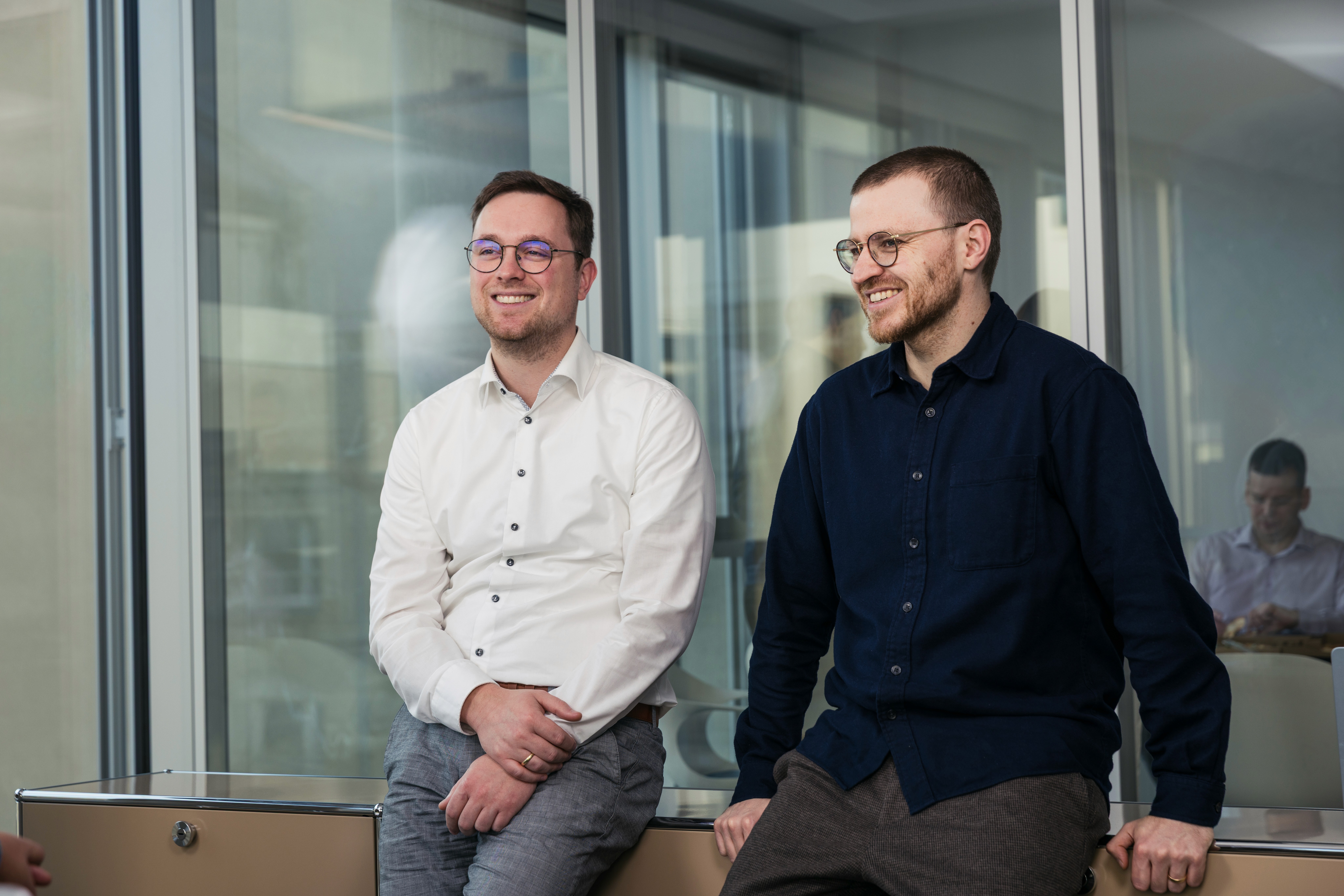 Zwei Mitglieder des Indicium Teams sitzen im Büro vor einer Fensterfront und lächeln in die Kamera