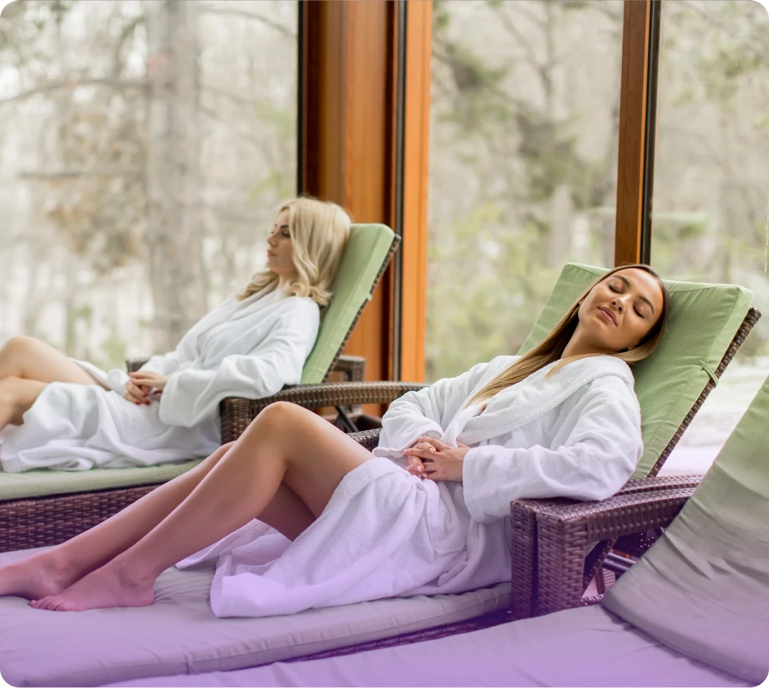 Two women in white robes relax on lounge chairs by large windows with natural light and a serene atmosphere.