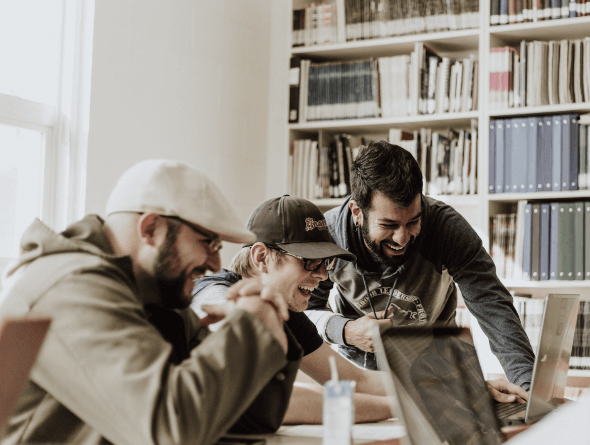 Three men gathered around a smartphone, engaged in conversation, with bookshelves in the background.