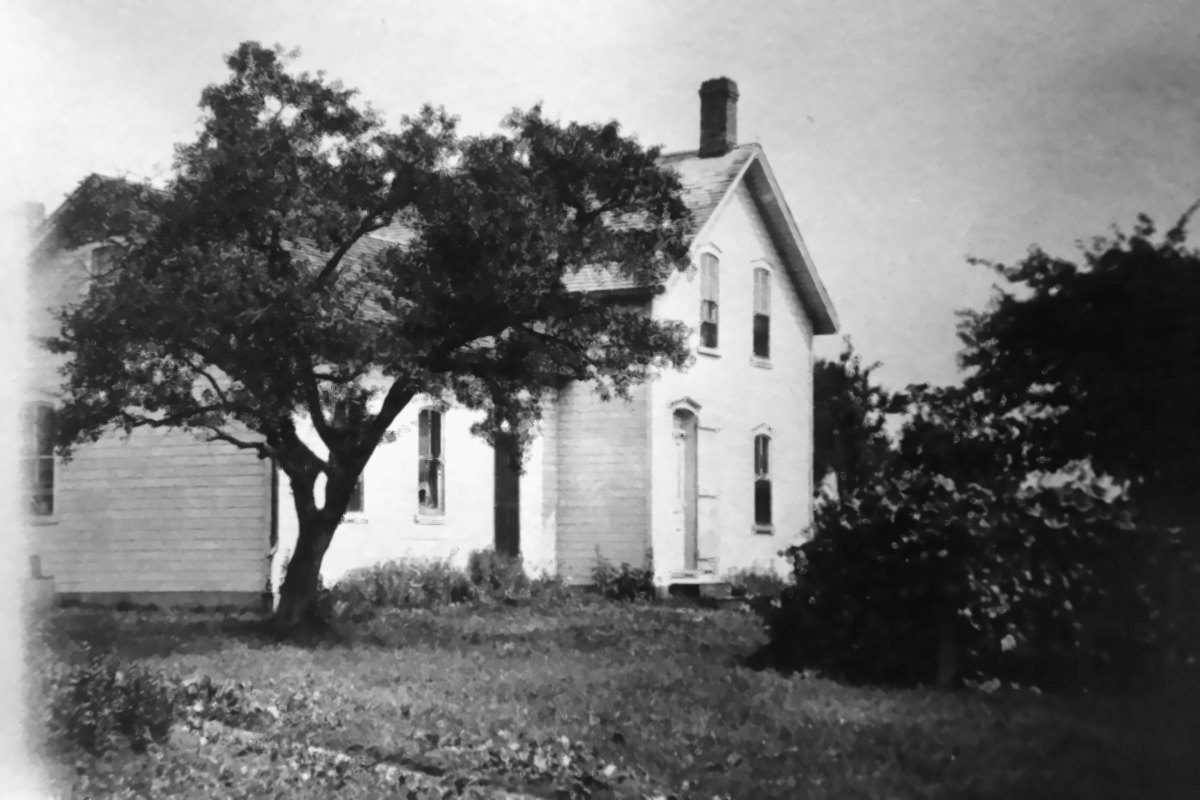 Restored black-and-white photograph of the original two-storey white clapboard Pfeifer farmhouse behind a spreading tree