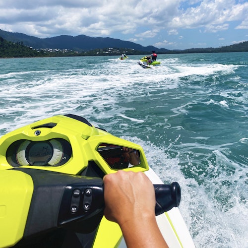 在半云的天空下，一名骑着黄色摩托艇的人在开阔水面上，追随远处的两艘摩托艇。