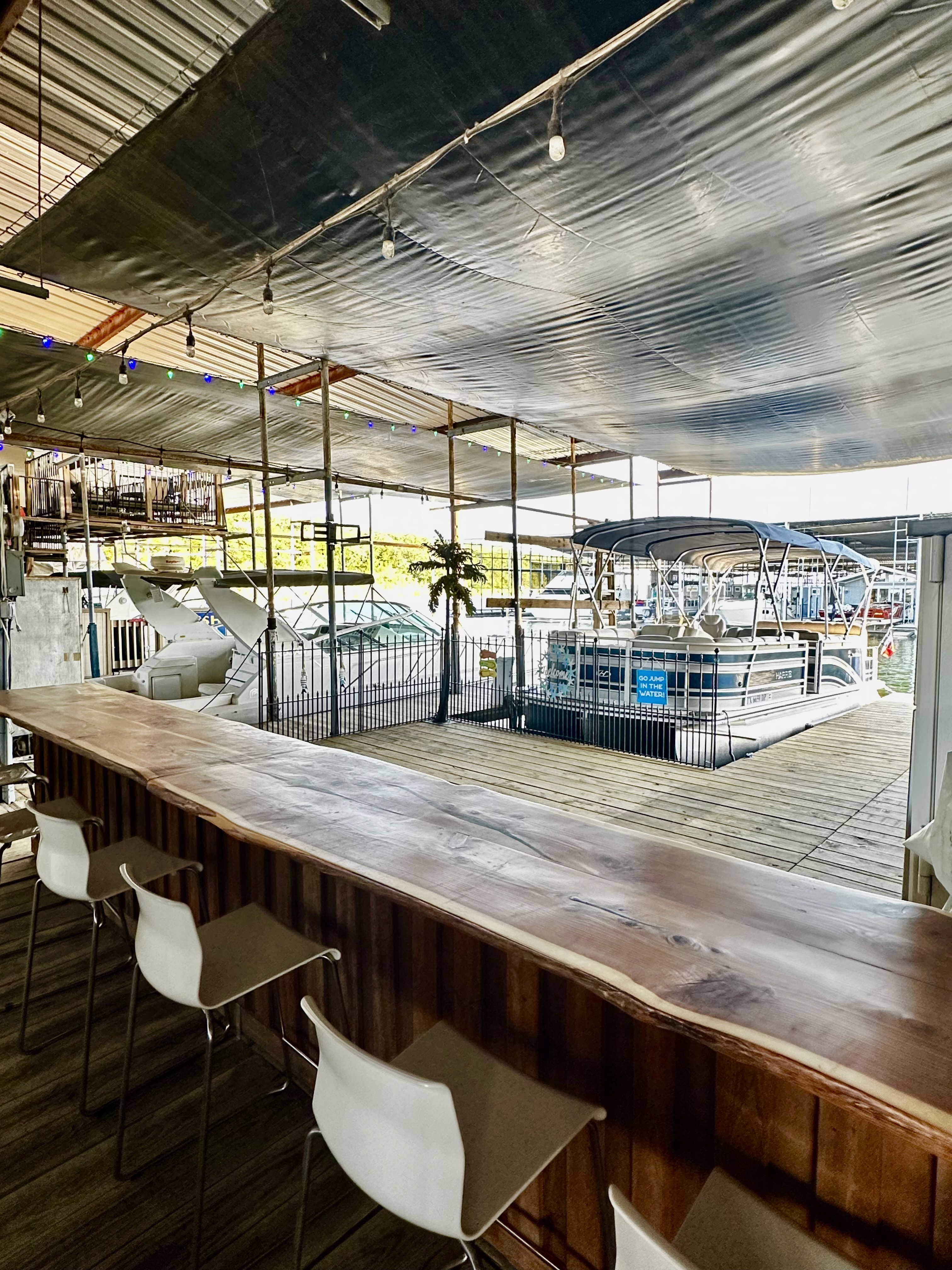 A spacious outdoor bar area with a long wooden counter, modern white barstools, and a view of boats docked under a metallic canopy, set in a nautical-themed environment with ambient string lighting.