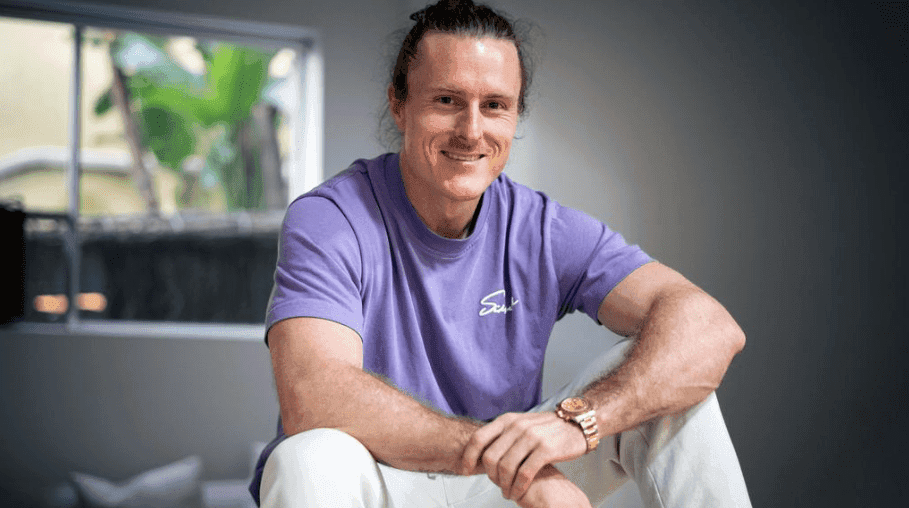 Man in purple t-shirt and white pants smiling.