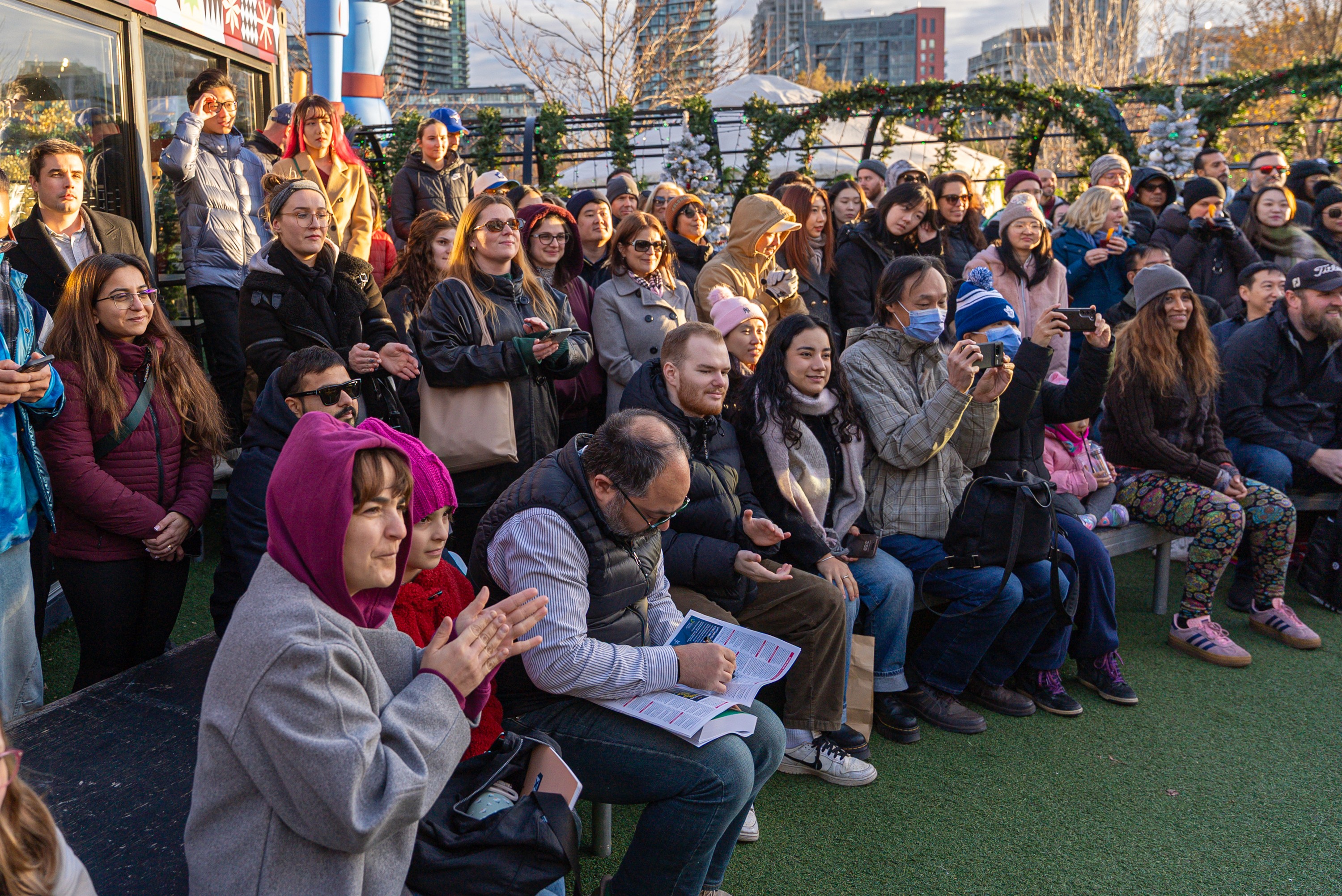 A large crowd gathered outdoors at the Taste of Iceland event in Toronto, watching a live presentation. People of all ages sit and stand closely together, many smiling, clapping, and taking photos in the winter sunlight.