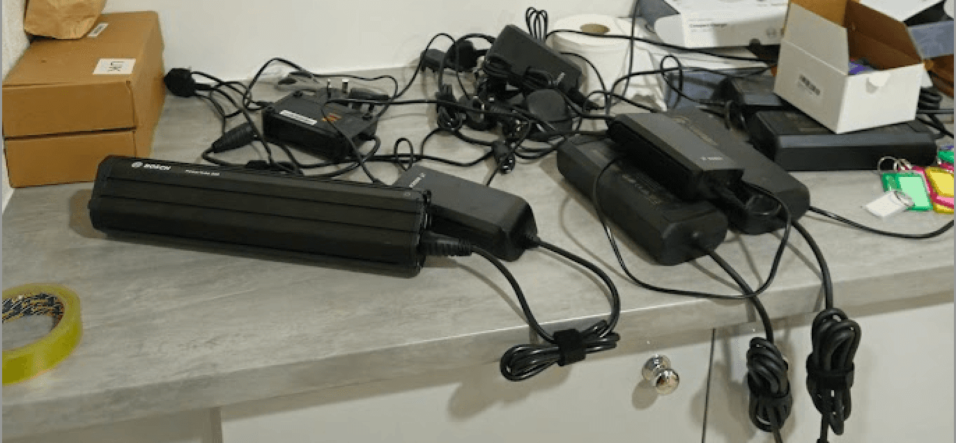 A real-world example of an unsafe e-bike charging setup on a counter, with tangled cables, power bricks, and flammable cardboard clutter nearby, illustrating a significant fire hazard that violates insurance policies
