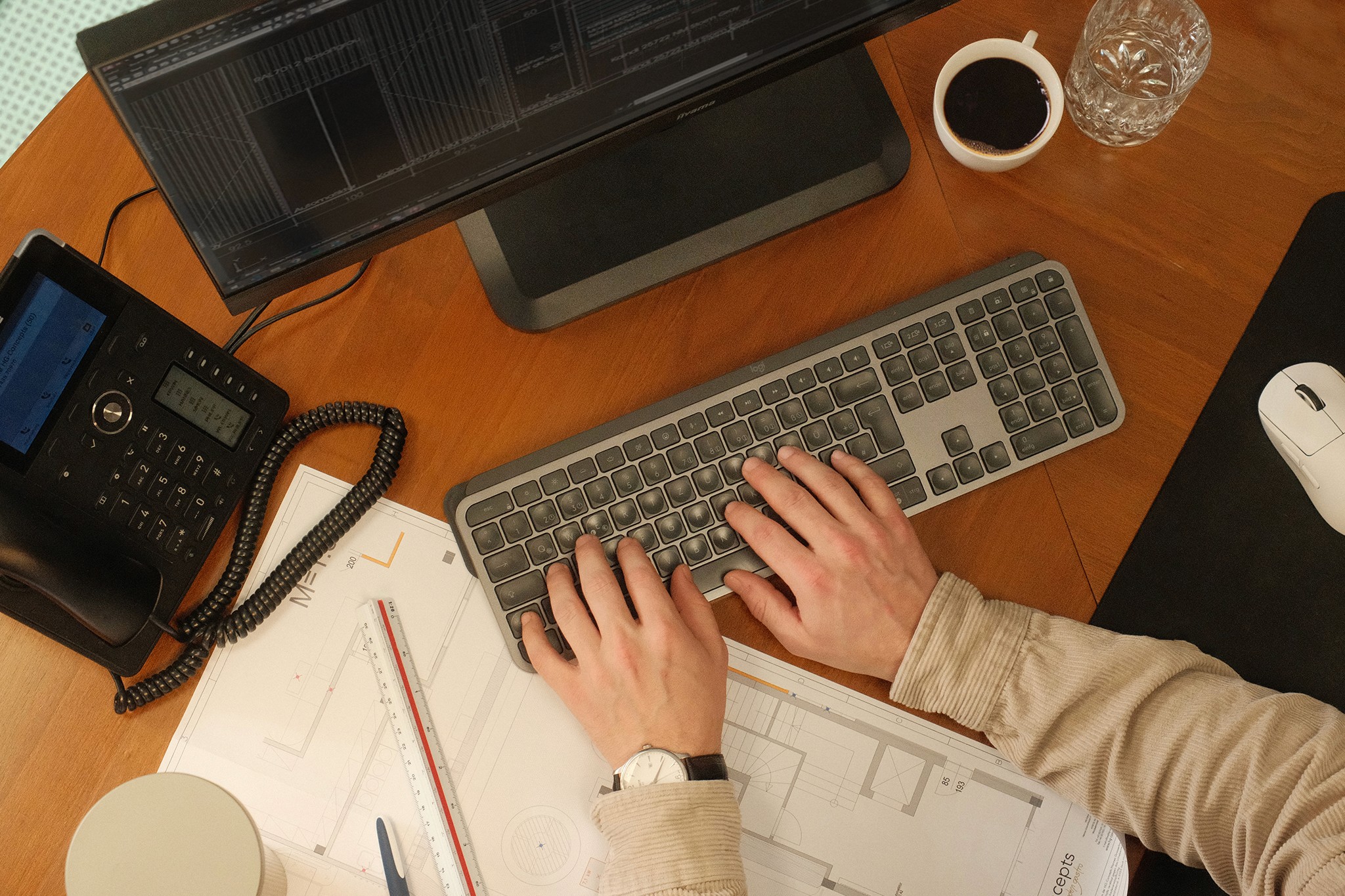 Detailbild einer Person die am Schreibtisch sitzt und arbeitet, mit Telefon, Plan und Tastatur