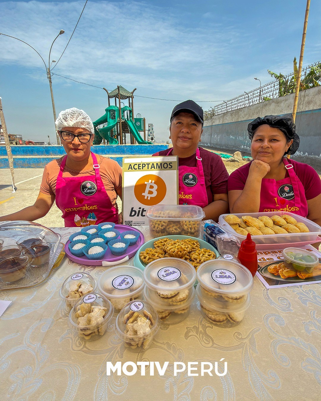 Lucía, Lidia, and Darlia — three food vendors wearing pink "I Love Cupcakes" aprons — stand behind an outdoor stall of homemade baked goods and snacks in Peru, with an "Aceptamos Bitcoin" sign displayed prominently on their table.