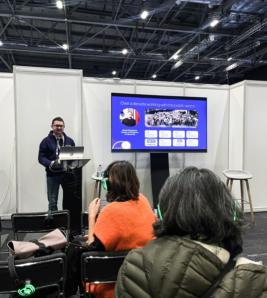 A presenter stands at a podium in a conference booth area, speaking to a small seated audience. A large screen beside him displays a slide about his decade of work in the public sector. Audience members wear green‑lit wireless headsets, and the venue’s high industrial ceiling is visible above the white partition walls.