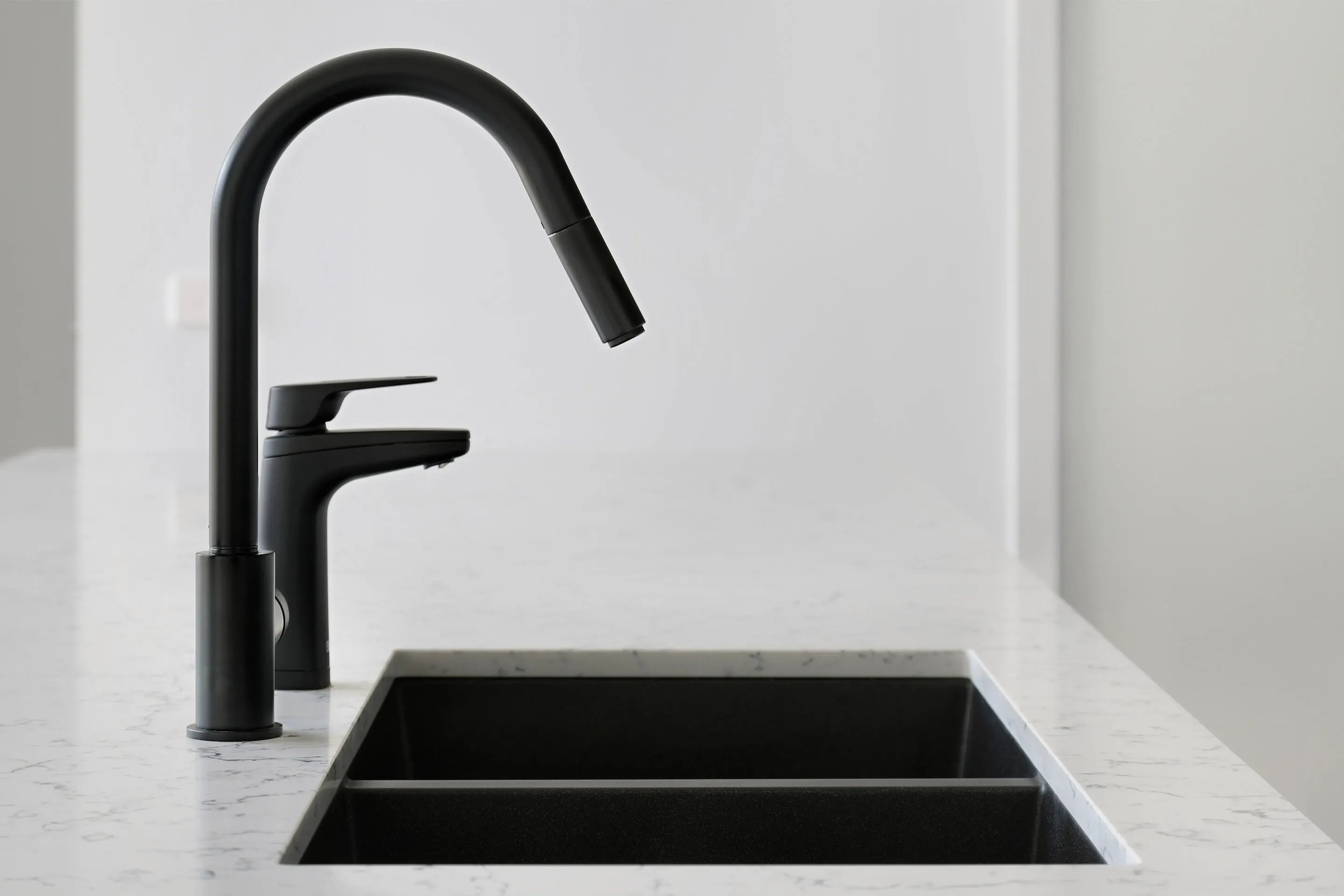 Kitchen detail of a matte-black gooseneck pull-down tap and mixer over a black undermount sink in a white veined stone benchtop