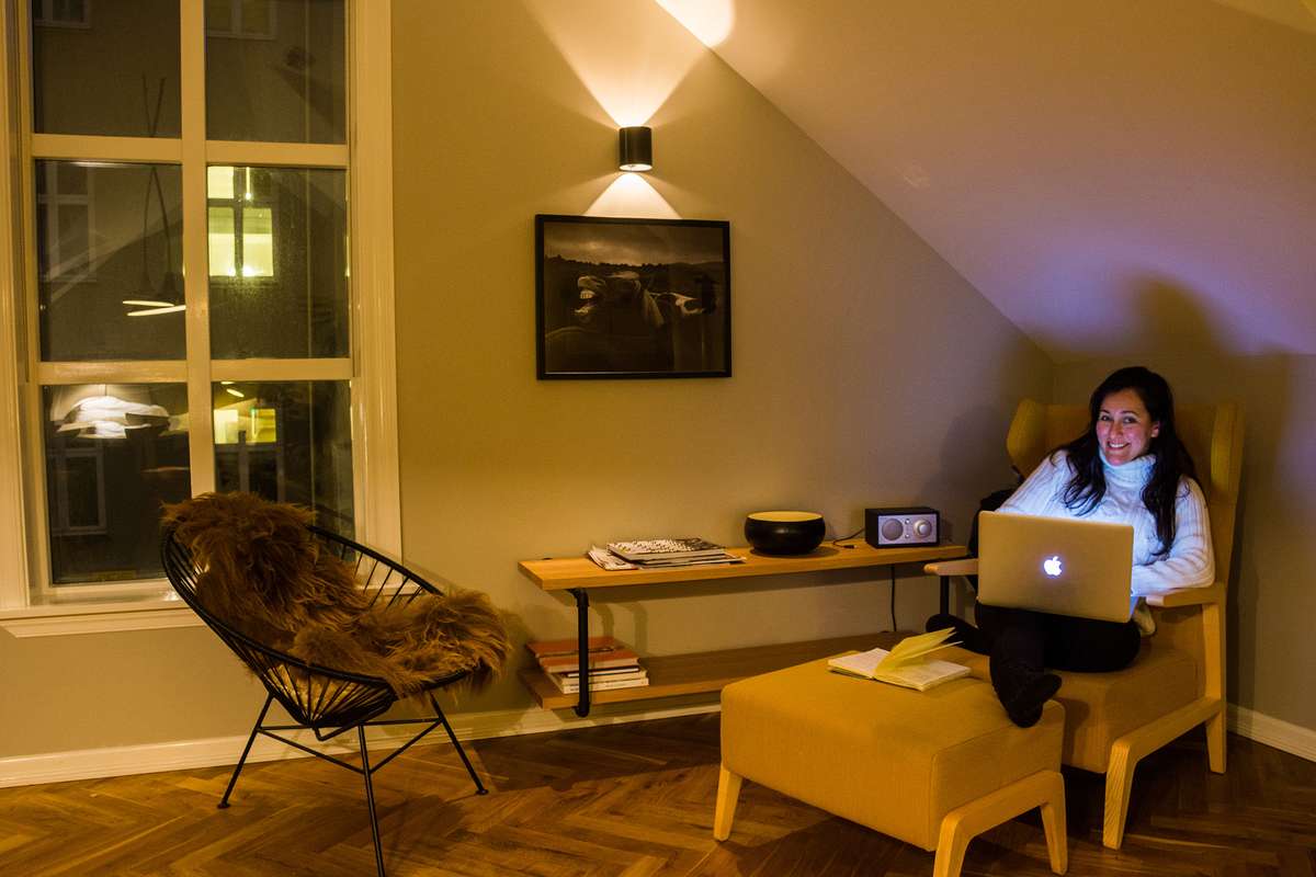 A woman sits in a yellow armchair in a cozy attic room with a laptop, a desk, and a chair draped in fur by a window.