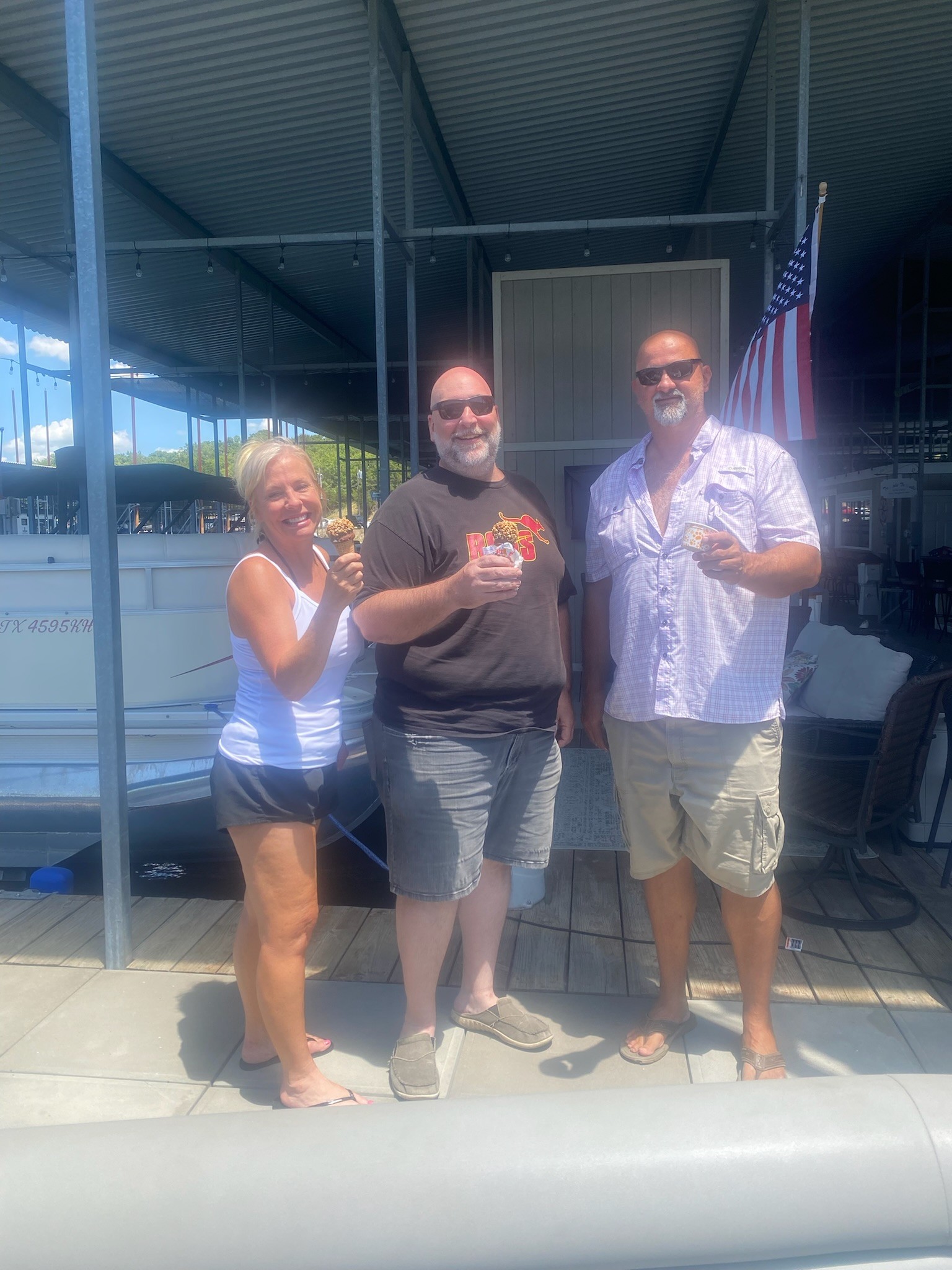 Three people stand smiling on a wooden dock near a boat, holding drinks and enjoying a sunny day under a shaded metal roof, with an American flag gracefully hanging in the background.