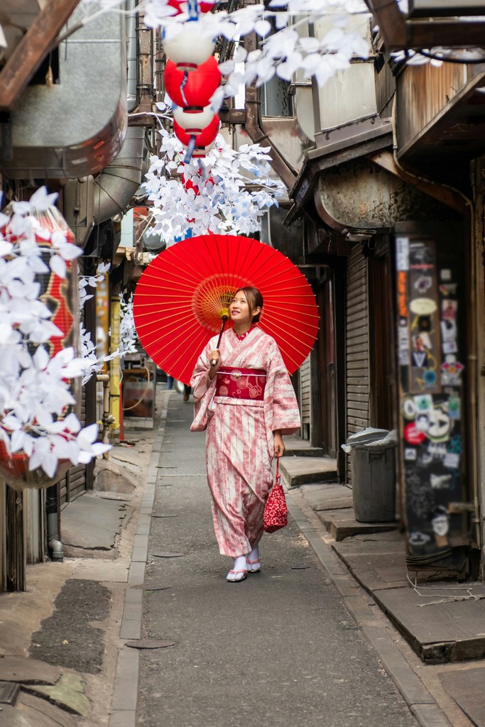 A woman in a traditional pink kimono walks down a narrow alleyway adorned with white paper decorations and red lanterns, holding a matching red parasol, embodying the elegance and cultural heritage of Japan.