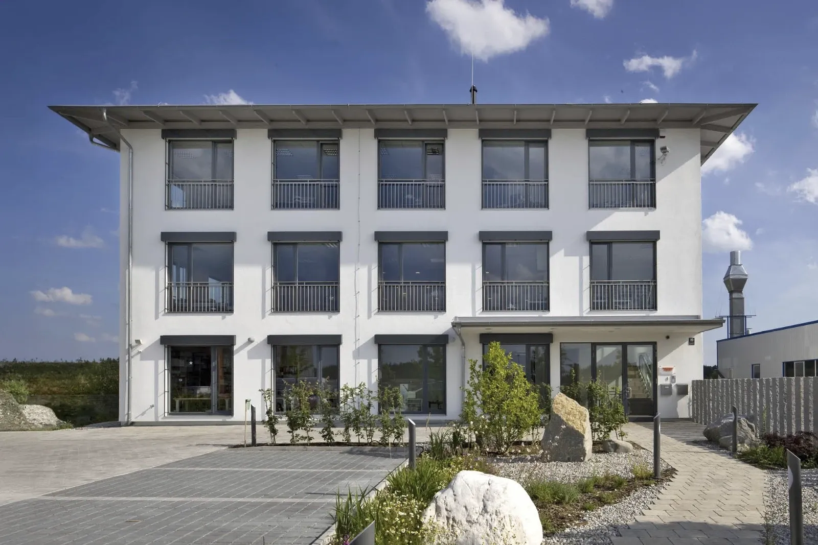 Dreistöckiges, weißes Bürogebäude mit großen Fenstern, Balkonen und einem flachen Dach, umgeben von Grünflächen und einem Parkplatz.