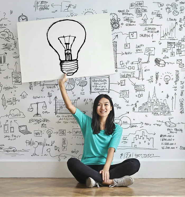 Woman sitting in front of ideas written on a wall, holding a drawing of a lightbulb.