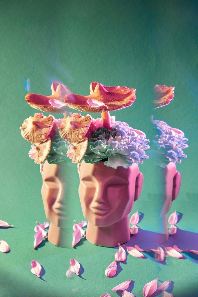 A surreal image features a ceramic head with mushrooms and flowers growing from the top, against a green background with scattered pink petals, symbolizing creativity, imagination, and the fusion of nature and art.