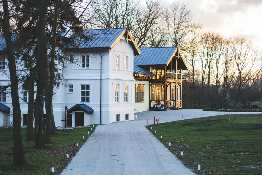 A large modern farmhouse-style home with white siding, black roof, and wraparound porch sits at the end of a long driveway lined with lights. The image represents luxury properties that qualify for Jumbo Loans from Chris Lewis Home Loans.