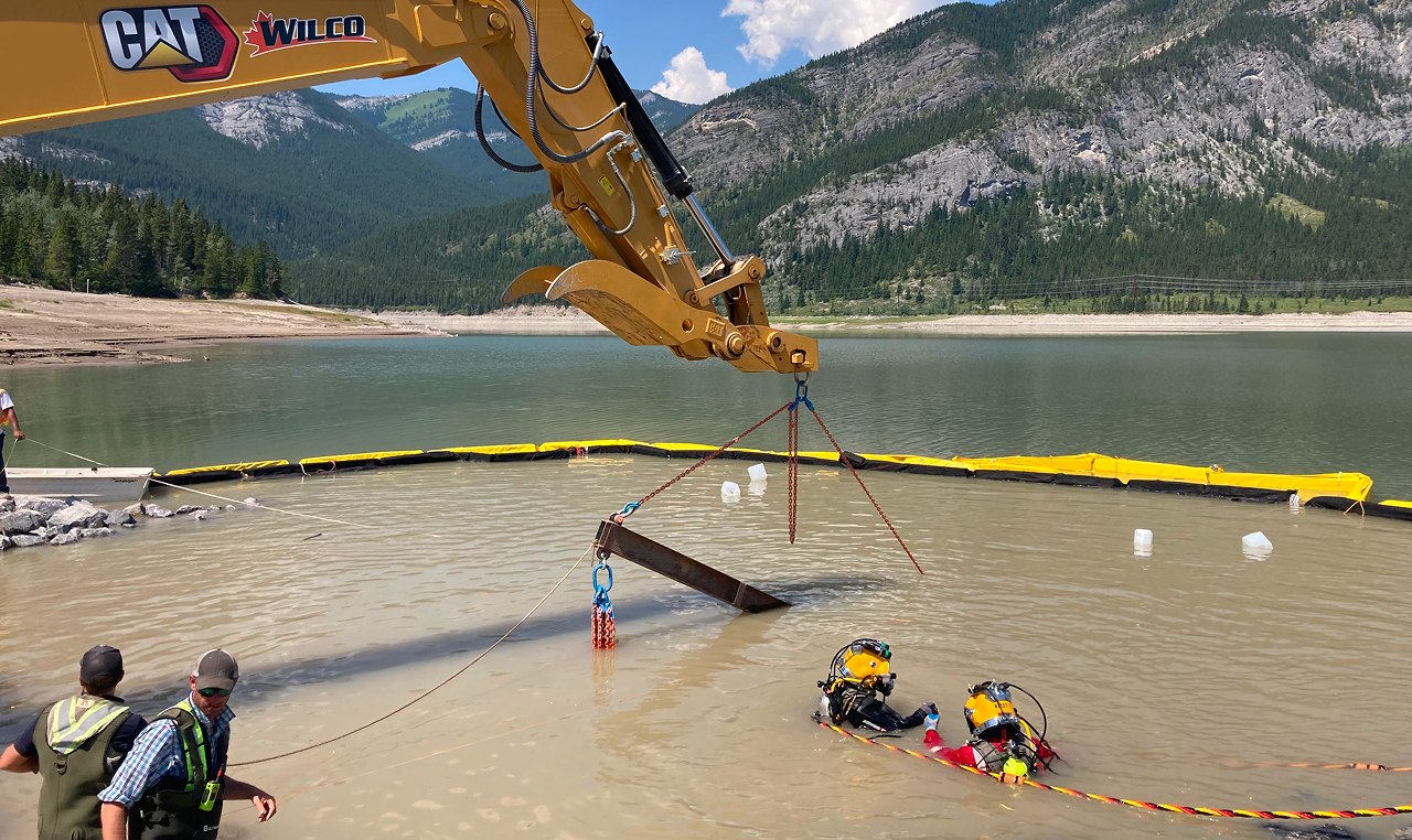 Construction crew using excavator to place structural beam underwater with turbidity curtain at Barrier Lake