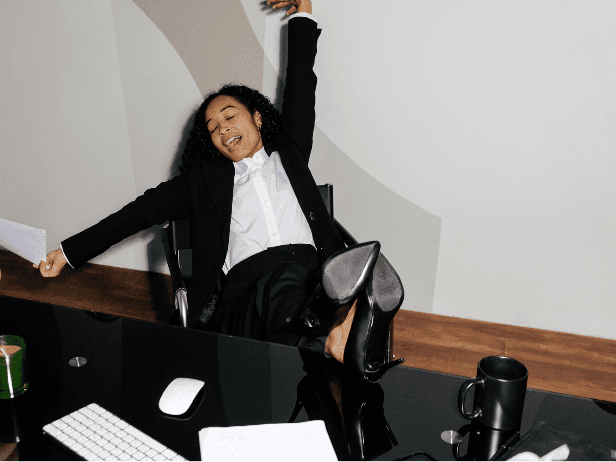 Joyful businesswoman celebrating at a desk with arms raised, sitting back in a modern office chair.