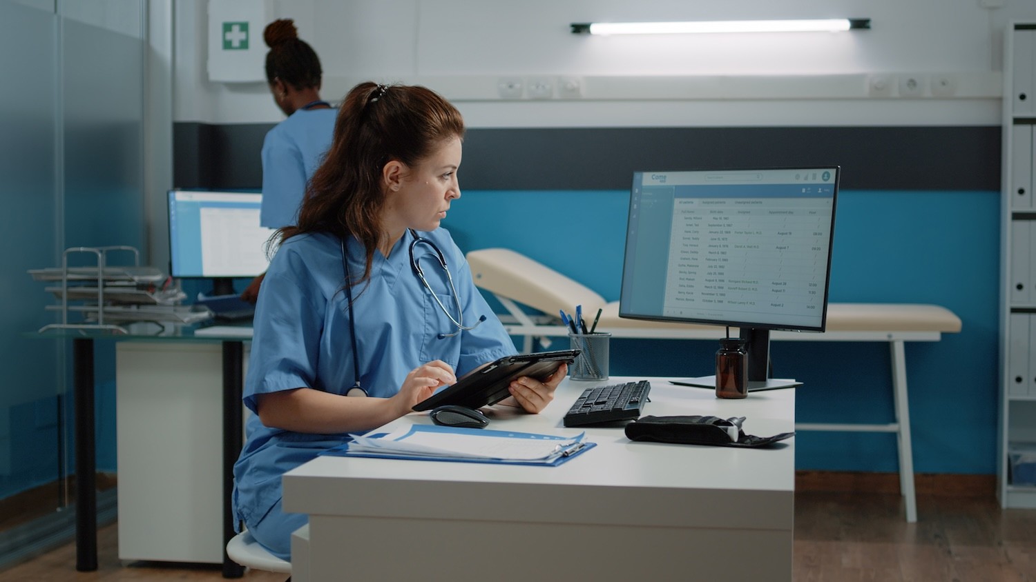 Eine Person in medizinischer Schutzkleidung sitzt an einem Schreibtisch und arbeitet mit einem Tablet vor einem Monitor in einem Krankenhauszimmer.