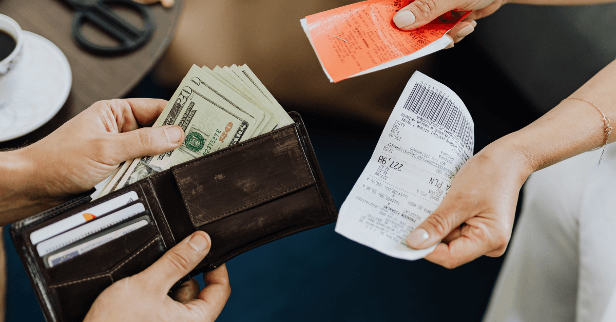 A closeup of two people's hands at a cafe. One person holds a receipt and the other holds an open wallet with cash and credit cards in it.