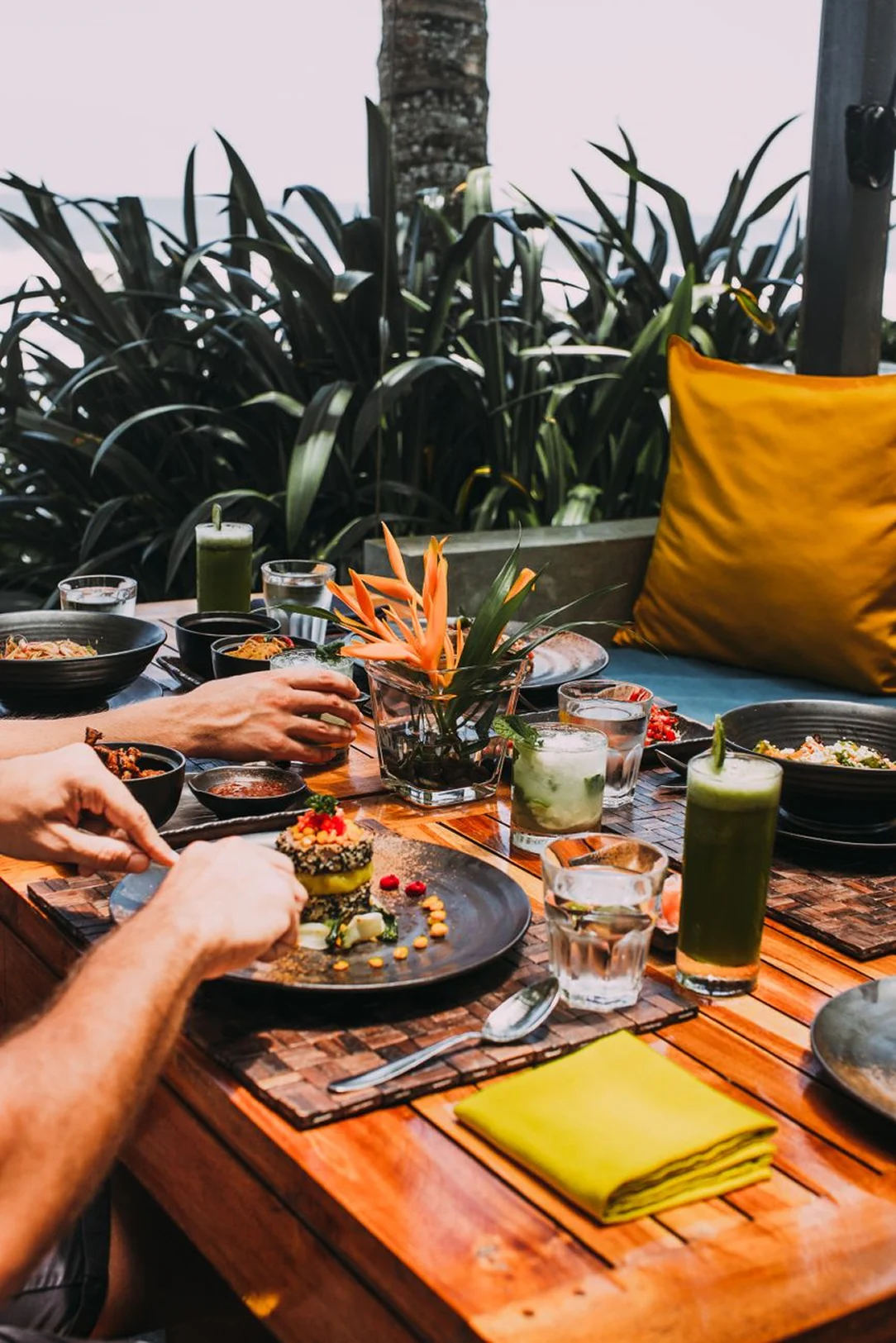 Guests sharing fresh dishes and drinks at an outdoor restaurant table surrounded by tropical plants.