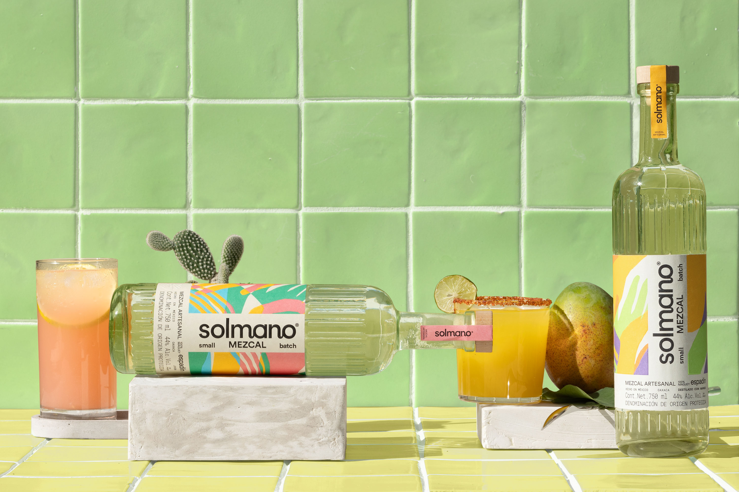 Solmano mezcal bottle and ready-to-serve cocktail cans displayed on marble beside fresh citrus and a pink cocktail against green tile