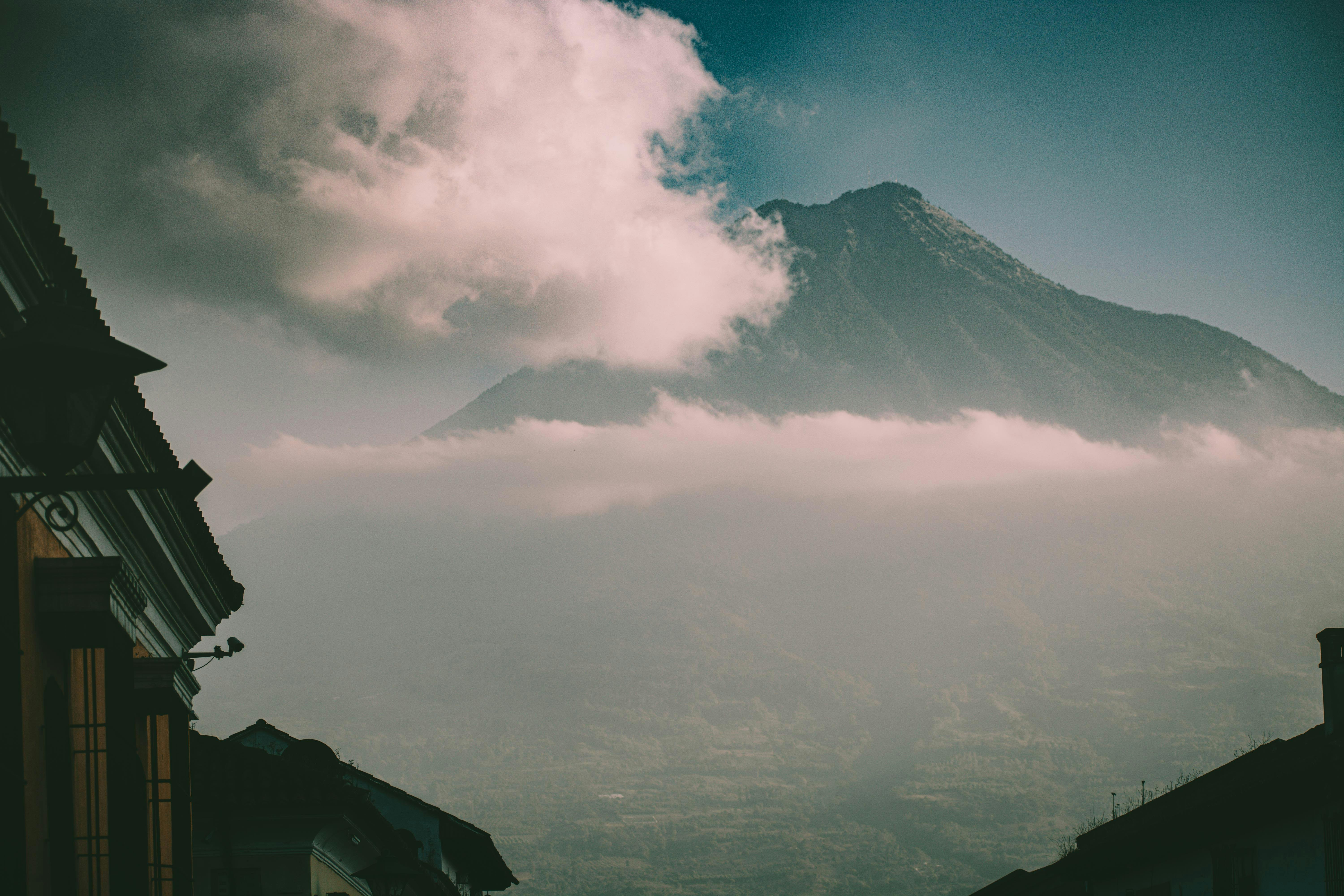 A dramatic view of a towering volcano, likely Volcán de Agua, partially obscured by soft white clouds. In the foreground, the dark silhouettes of colonial-style rooftops and a street lamp are visible against the hazy mountain backdrop.