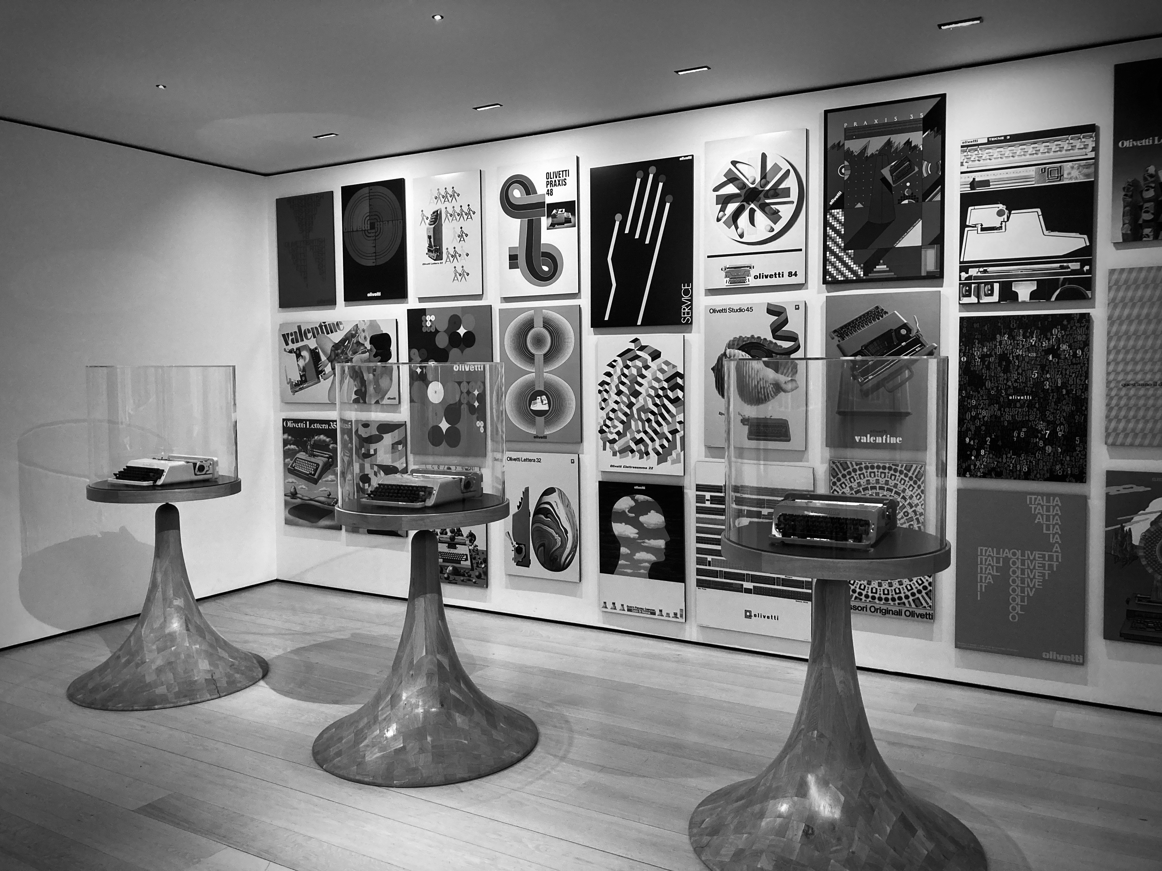 Black and white photo from a museum in London, showing three old typewriters and artistic posters on the wall in the background.
