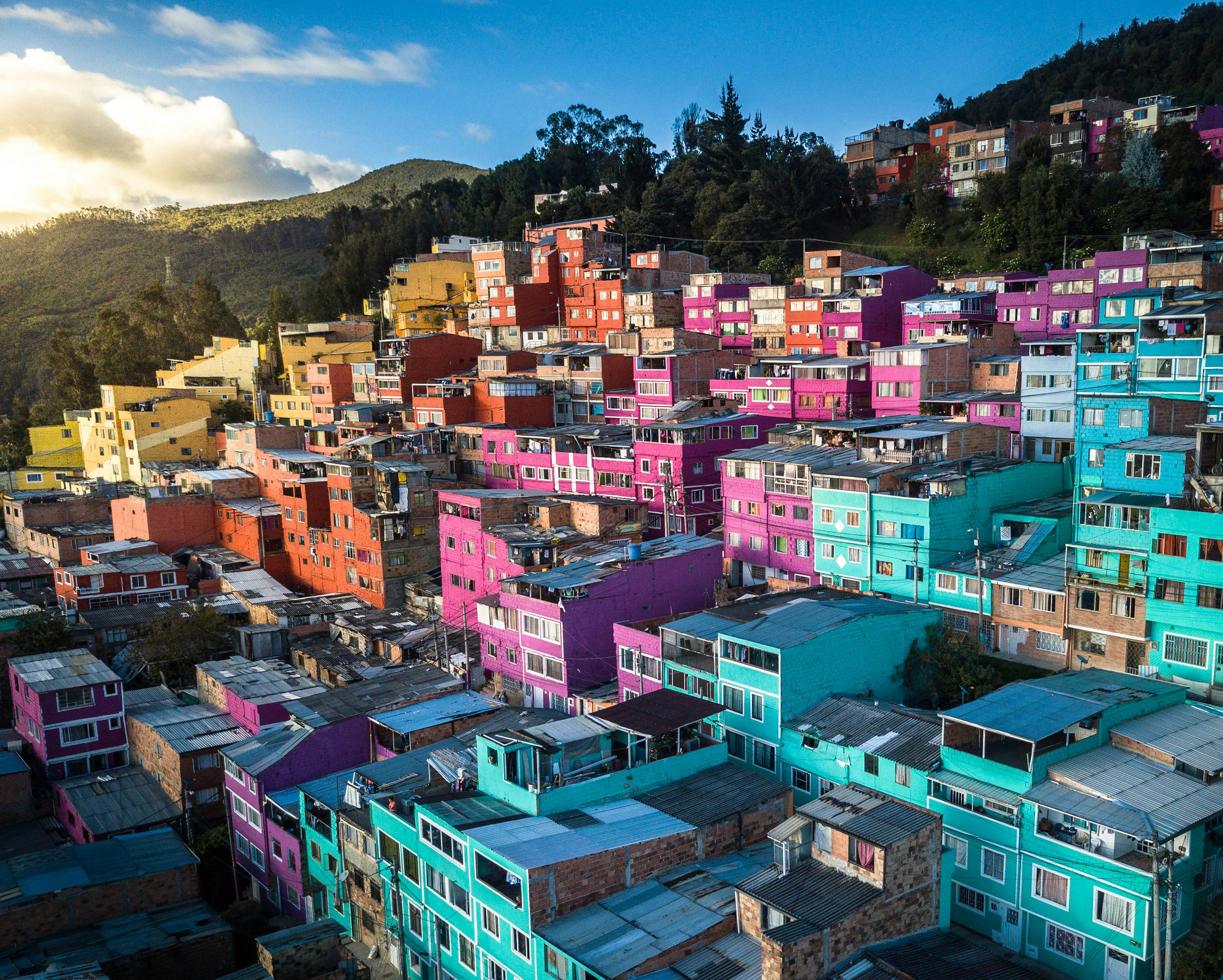 A hillside neighborhood in Quito featuring rows of densely packed houses painted in vivid shades of purple, teal, pink, and orange. The morning sun hits the side of the mountain, creating a bright contrast against the colorful urban landscape.