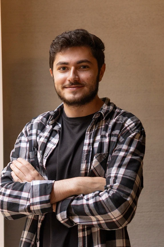 Half-length portrait in a black tee and plaid shirt, arms crossed, neutral wall background.