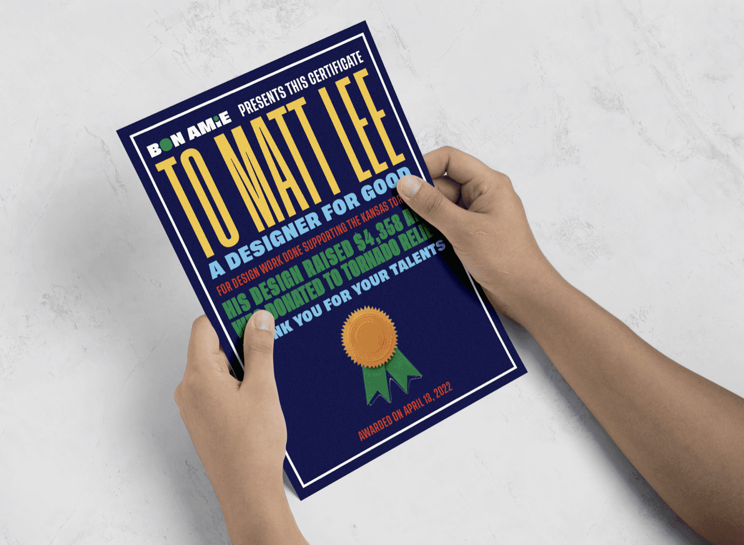 Two hands hold a dark blue book titled “TO MAT TEE” with yellow and blue text. The subtitle reads “A DESIGNER FOR GOOD.” Below it, a smaller text states, “FOR WORK DONE SUPPORTING THE KANSAS TOUR HAS BEEN RENOWNED TO TORCHED LELO.” A gold medal graphic is centered on the cover. The book is marked “AWARDED ON APRIL 13, 2022.”