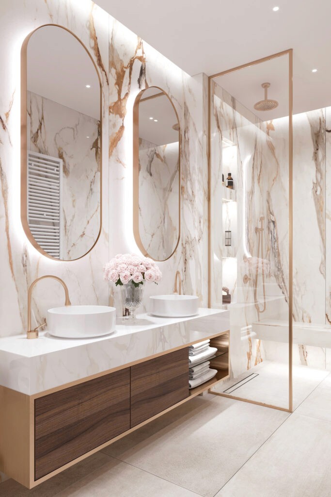 Modern bathroom with marble walls and countertops, two oval mirrors, dual white vessel sinks with gold faucets, and a vase of pink roses centered between the sinks