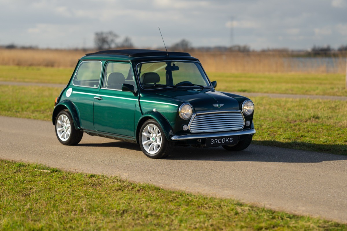 A classic green car parked on a rural road, with a grassy field and blue sky in the background.
