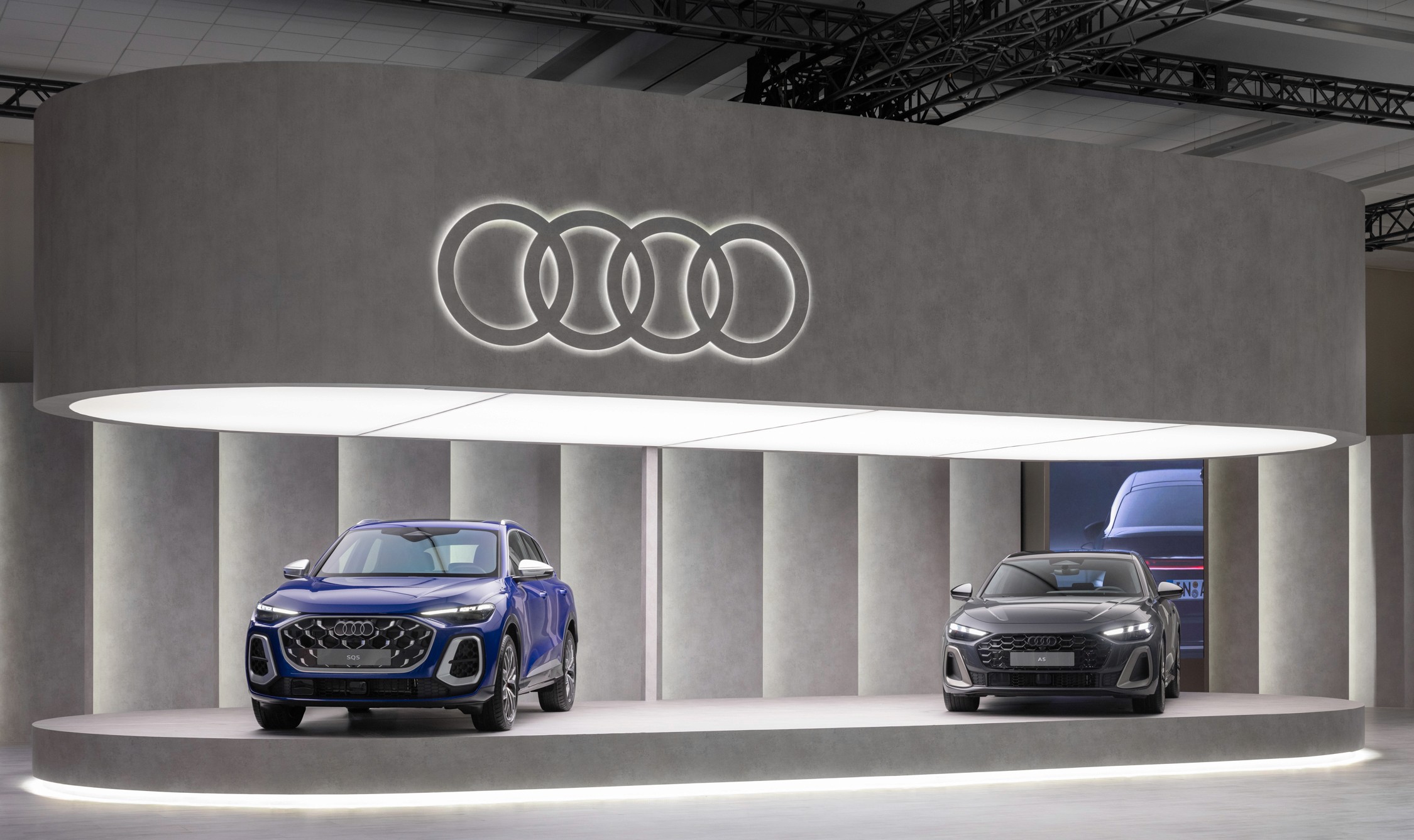 Two sleek Audi cars, one blue and one black, are displayed on a modern, illuminated platform with the Audi logo overhead, exuding luxury and sophistication.