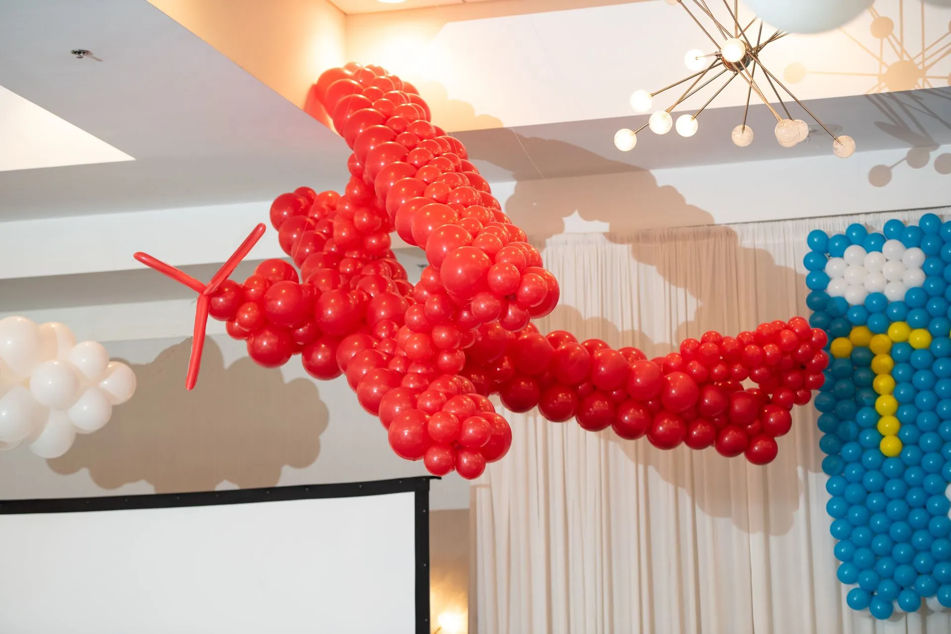 A vibrant red airplane sculpture made entirely of balloons is suspended in a room, with white balloon clouds and decorative lighting visible in the background, creating a festive atmosphere.